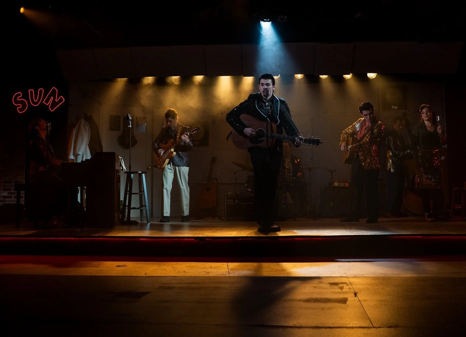 A band performing on a dimly lit stage, including a male singer playing guitar, a pianist, a guitarist, and three backup singers. The stage has warm yellow and blue lighting, with a neon red sign that says 'sun' in the background.