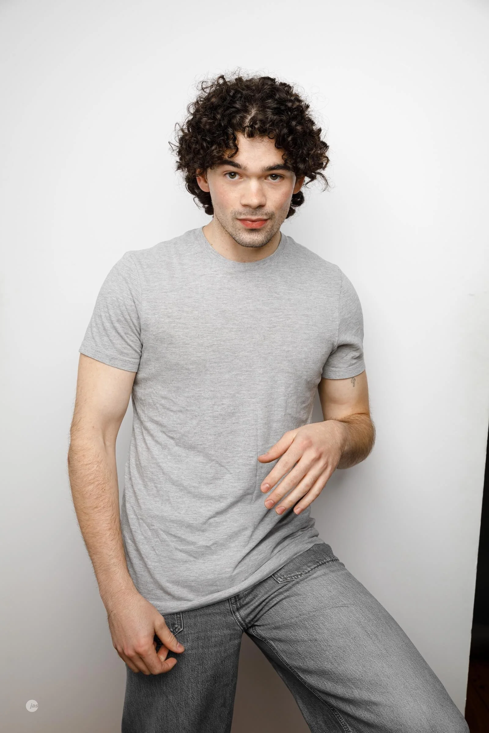 A young man with curly dark hair wearing a light grey t-shirt and grey jeans, standing against a white background.