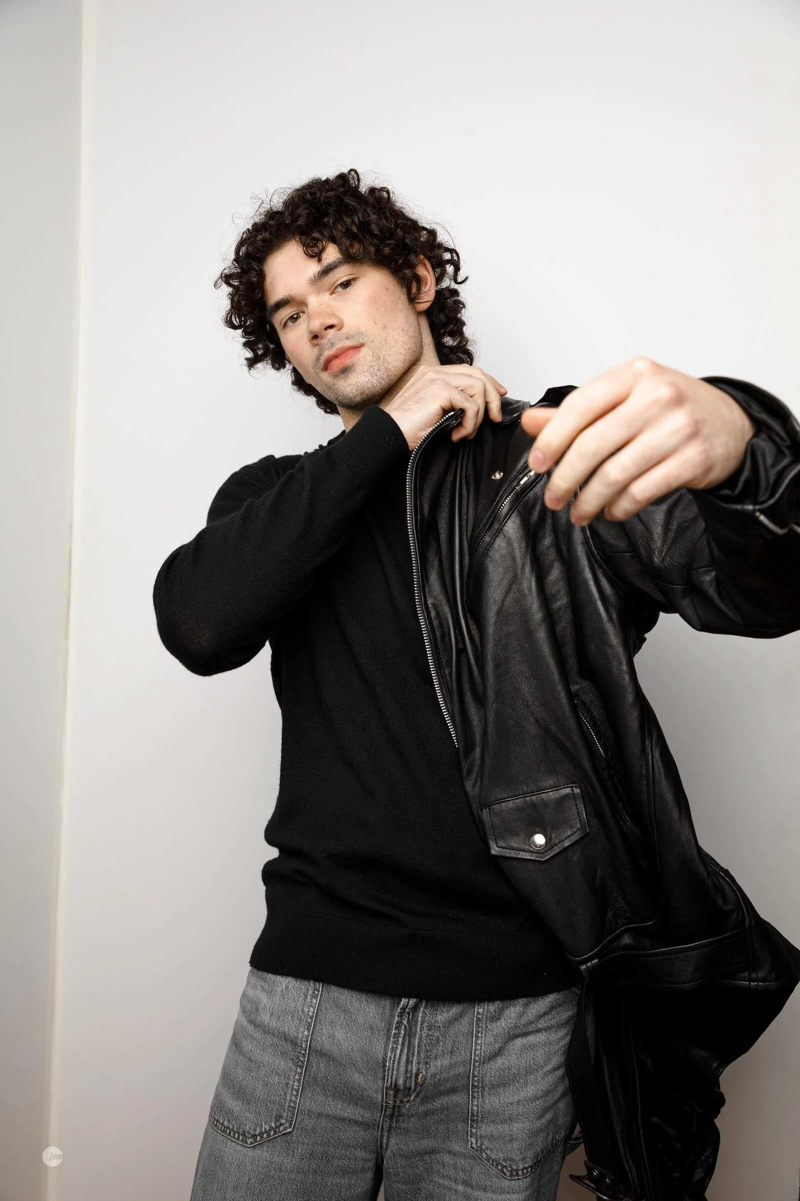 A young man with curly hair in a black shirt and gray jeans is putting on a black leather jacket against a plain white background.
