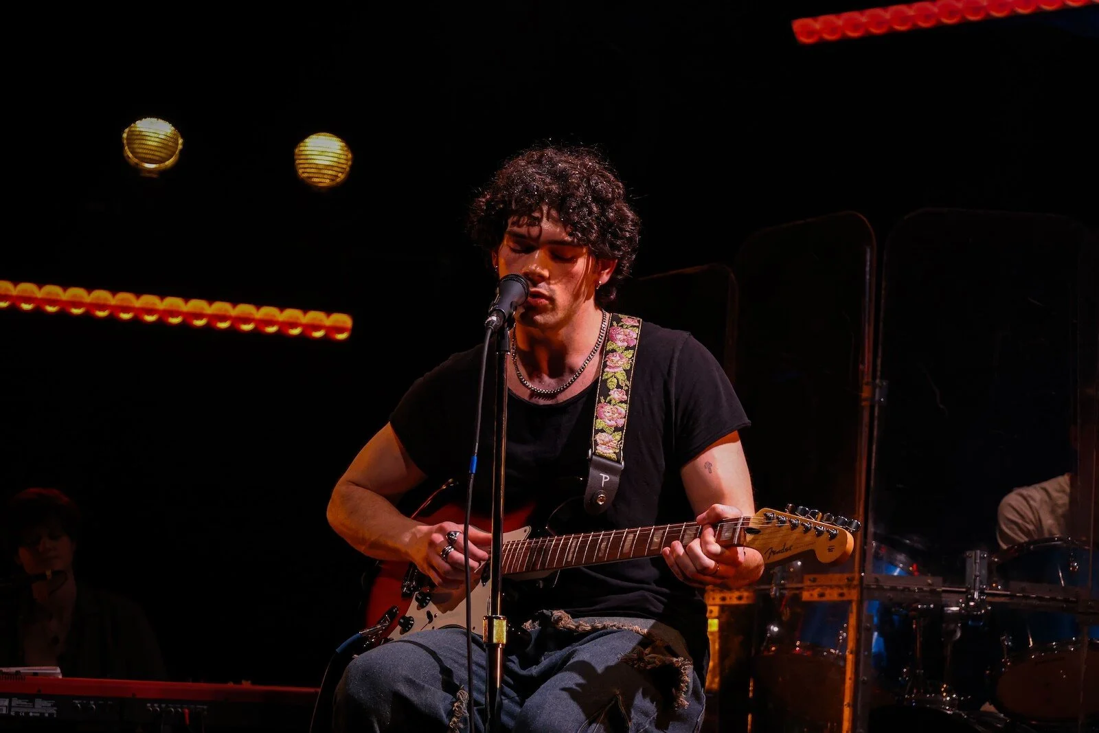 A young man with curly hair playing an electric guitar and singing into a microphone on stage, with a keyboard and other band members in the background.