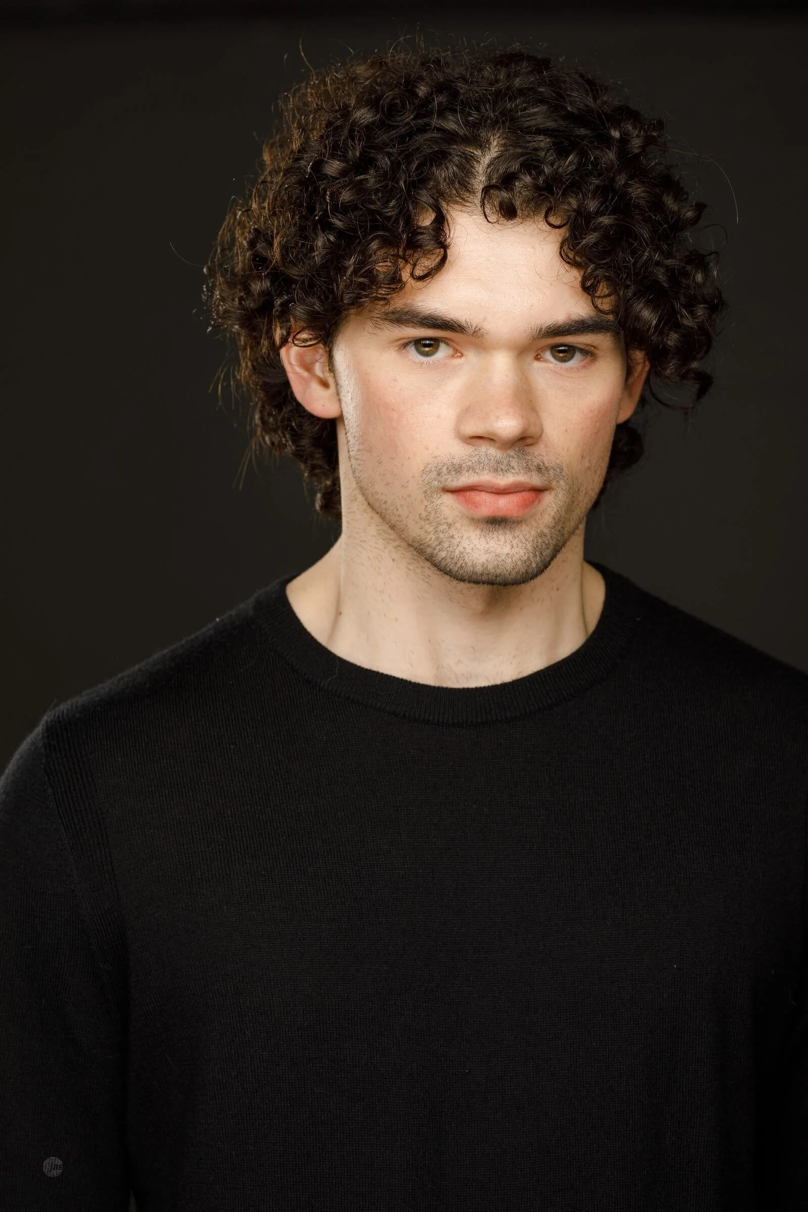Portrait of a young man with curly brown hair and light skin, wearing a black shirt against a dark background.