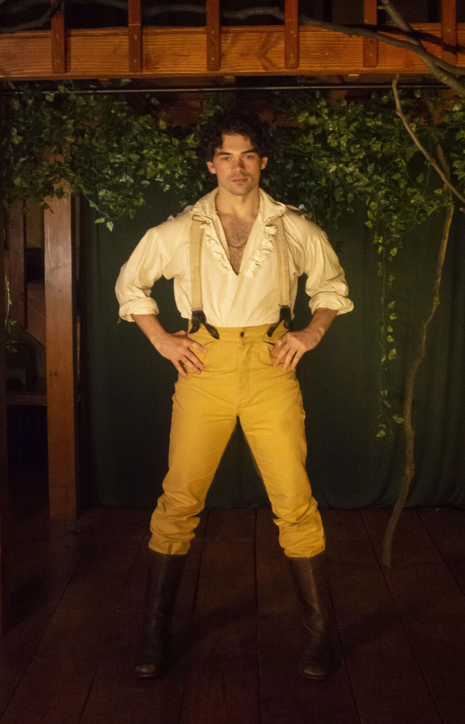 A man standing with hands on hips, wearing vintage yellow suspenders, beige shirt, yellow pants, and tall black boots, in a setting with wooden beams and greenery.