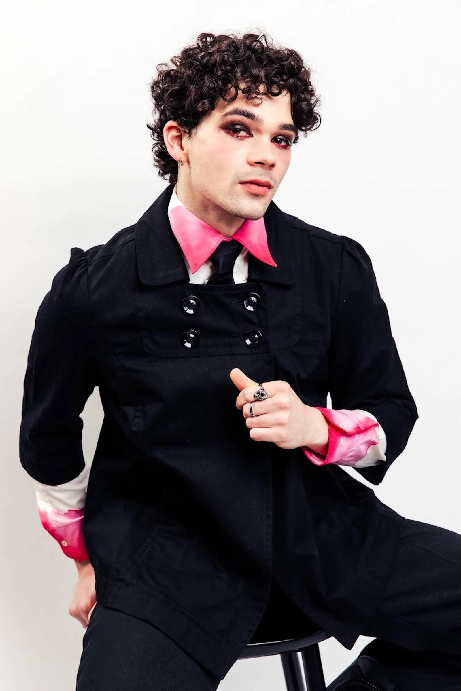 Person with curly hair wearing a black jacket with pink and white shirt underneath, seated on a stool, against a plain white background.