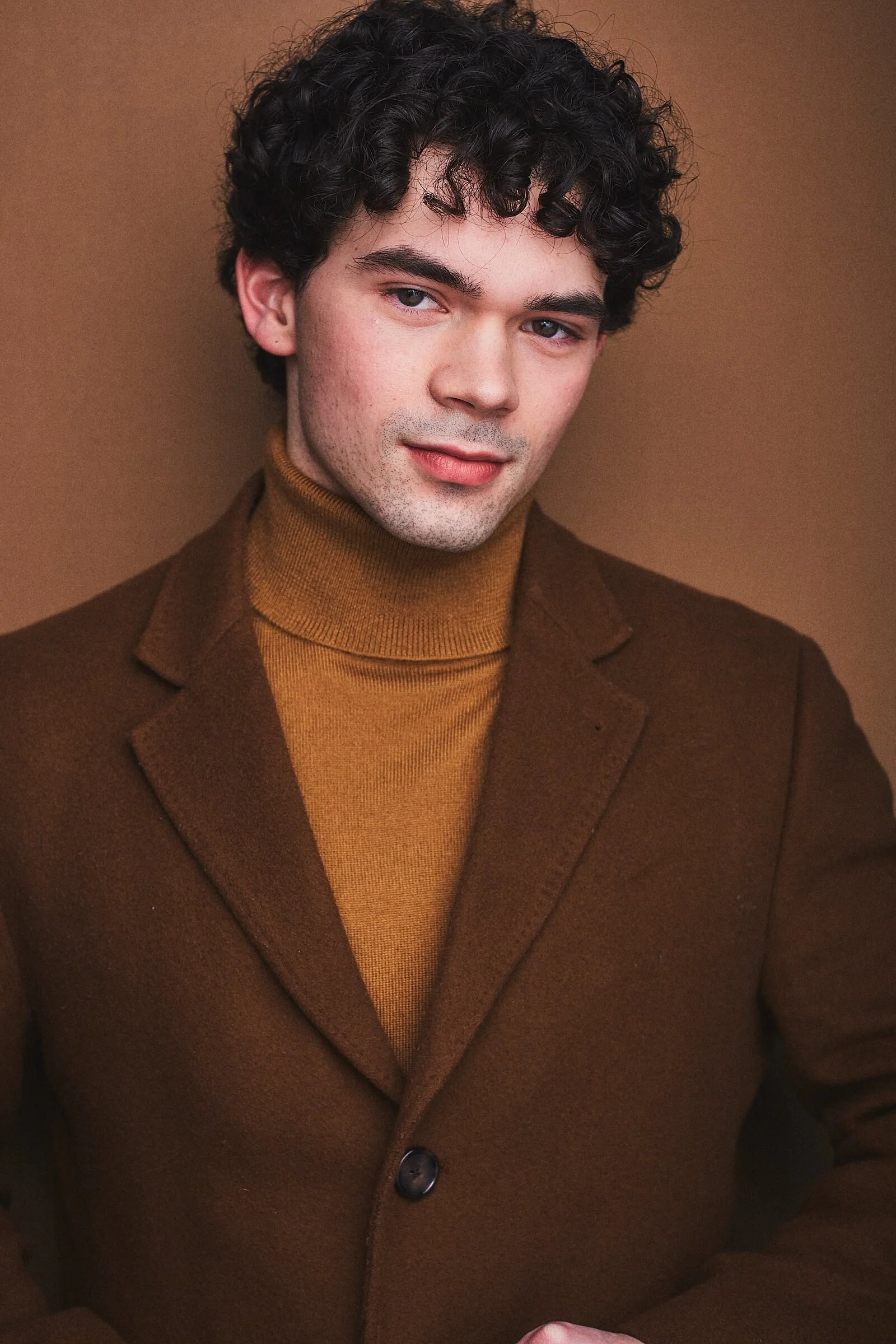 A young man with curly dark hair, light skin, and blue eyes, dressed in a brown jacket and a mustard turtleneck sweater, posing against a brown background.