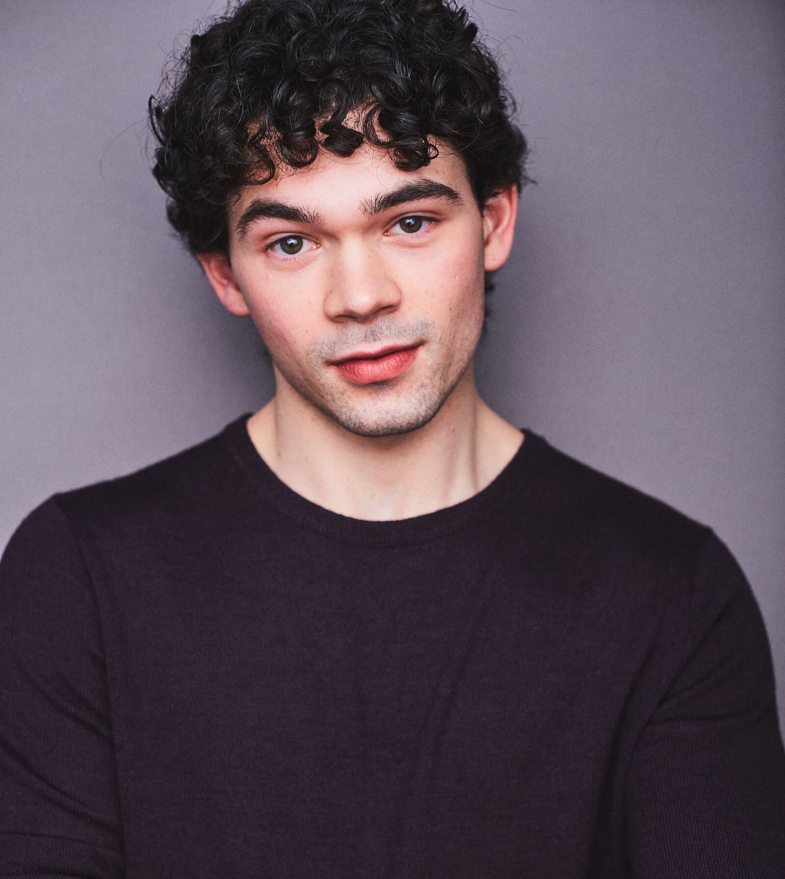 Portrait of a young man with curly dark hair wearing a black shirt, standing against a gray background