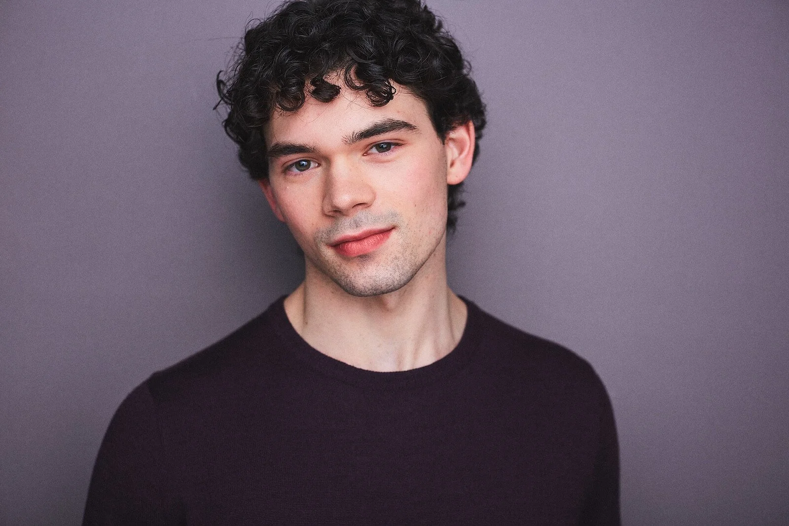 A young man with curly dark hair, blue eyes, and light skin, wearing a black shirt, posed against a plain gray background.