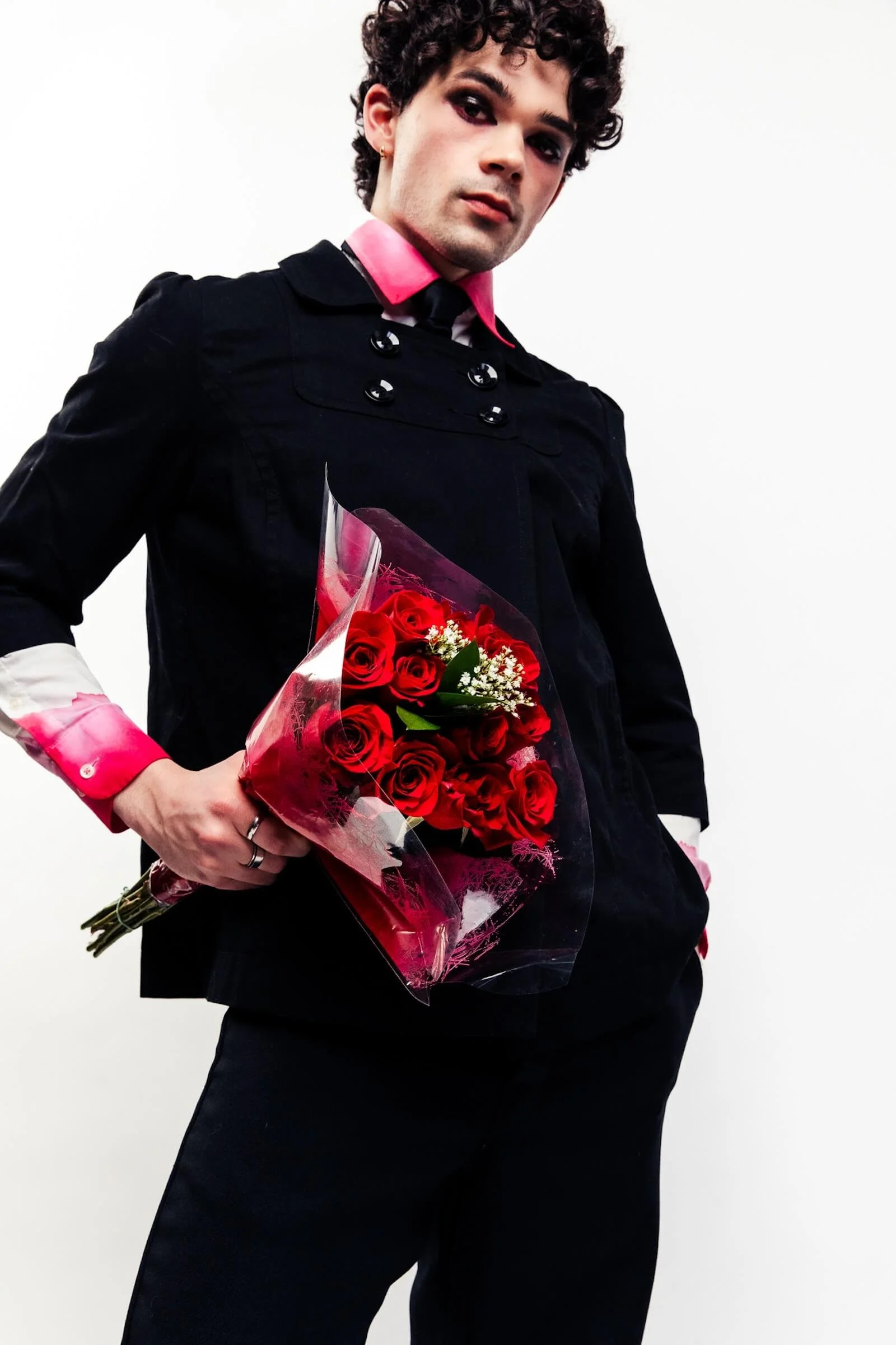 A person with dark curly hair wearing a black jacket with pink shirt cuffs and holding a bouquet of red roses.