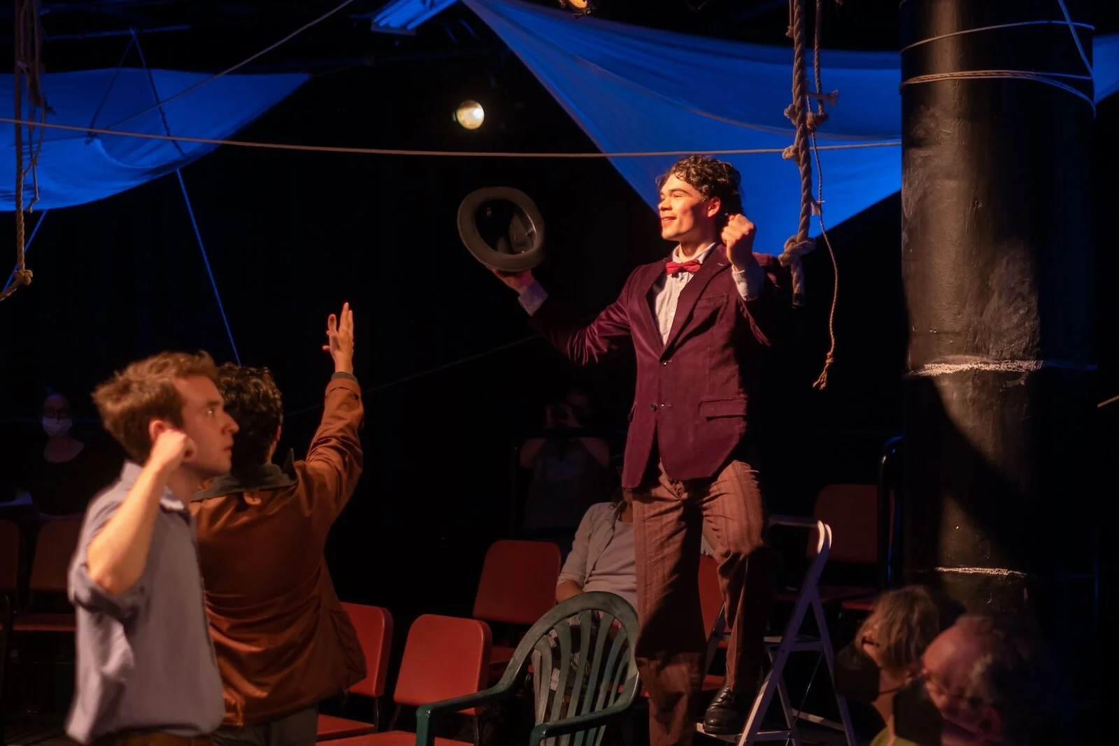 A theatrical performance with a young man in a burgundy jacket, bow tie, and striped pants, standing on a chair with a hat in hand, smiling, while engaging with two other actors raising their hands, in a dimly lit stage with blue props overhead.