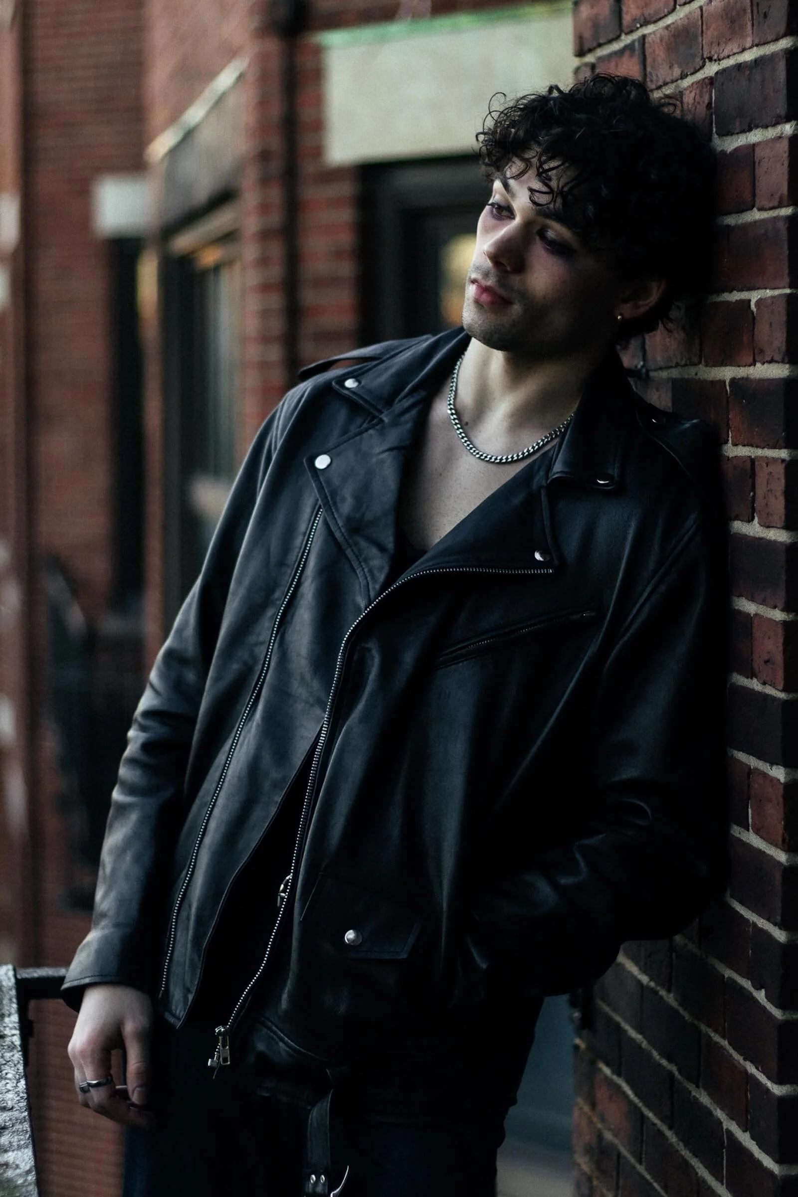 A young man with curly dark hair leaning against a brick wall, wearing a black leather jacket, a silver chain necklace, and rings.