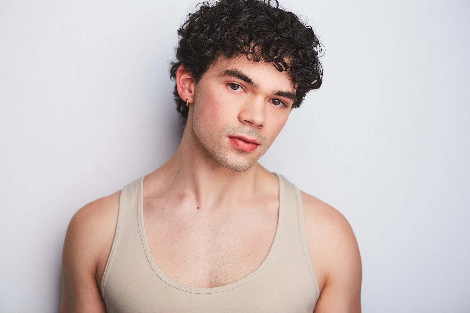 A young man with curly dark hair, light skin, and wearing a beige tank top, is standing against a plain white wall, looking directly at the camera.