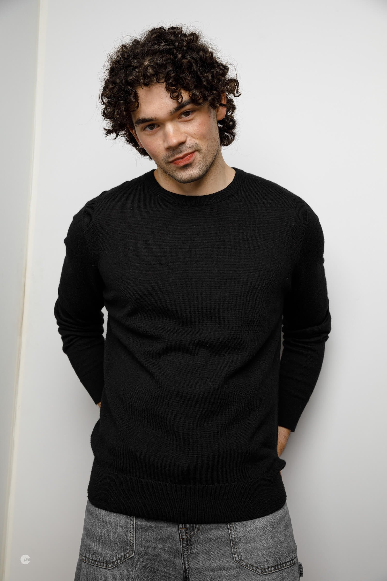 A young man with curly dark hair, wearing a black sweater and gray jeans, standing against a plain white background, looking at the camera with a slight smile.
