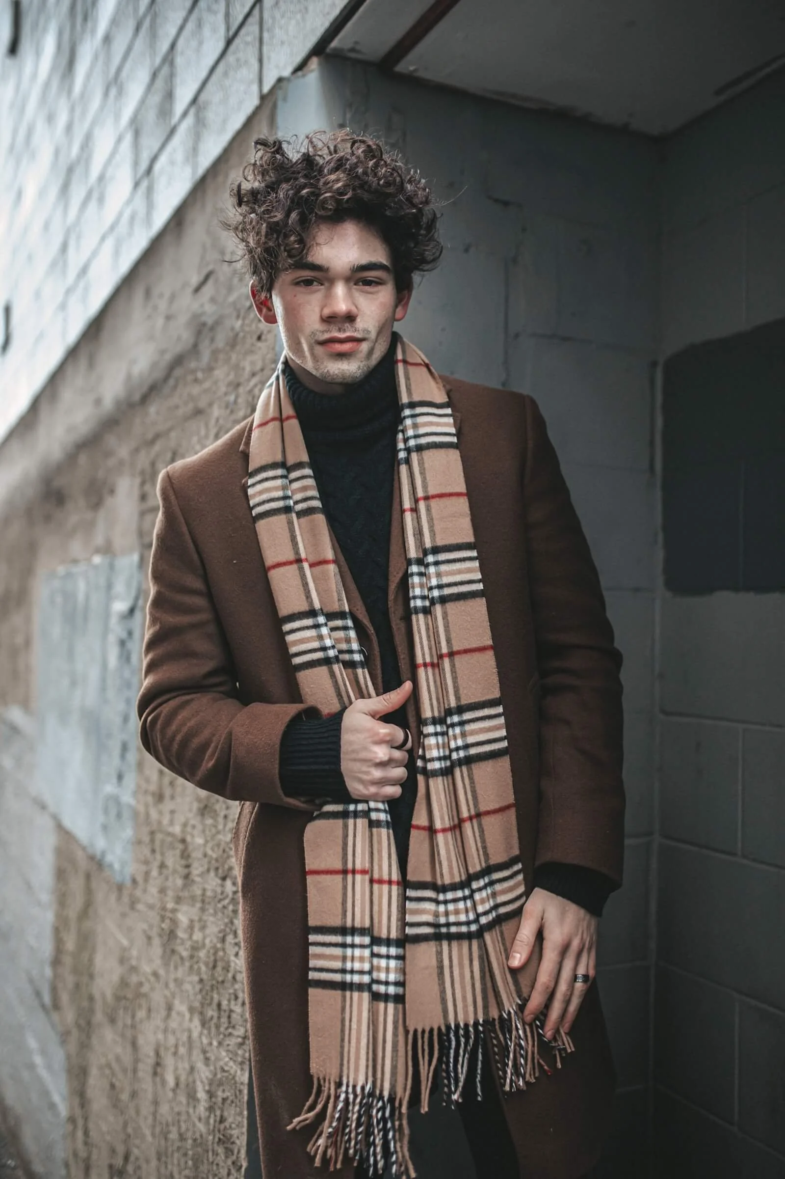 A young man with curly hair wearing a brown coat, black turtleneck, and beige plaid scarf standing against a concrete wall.