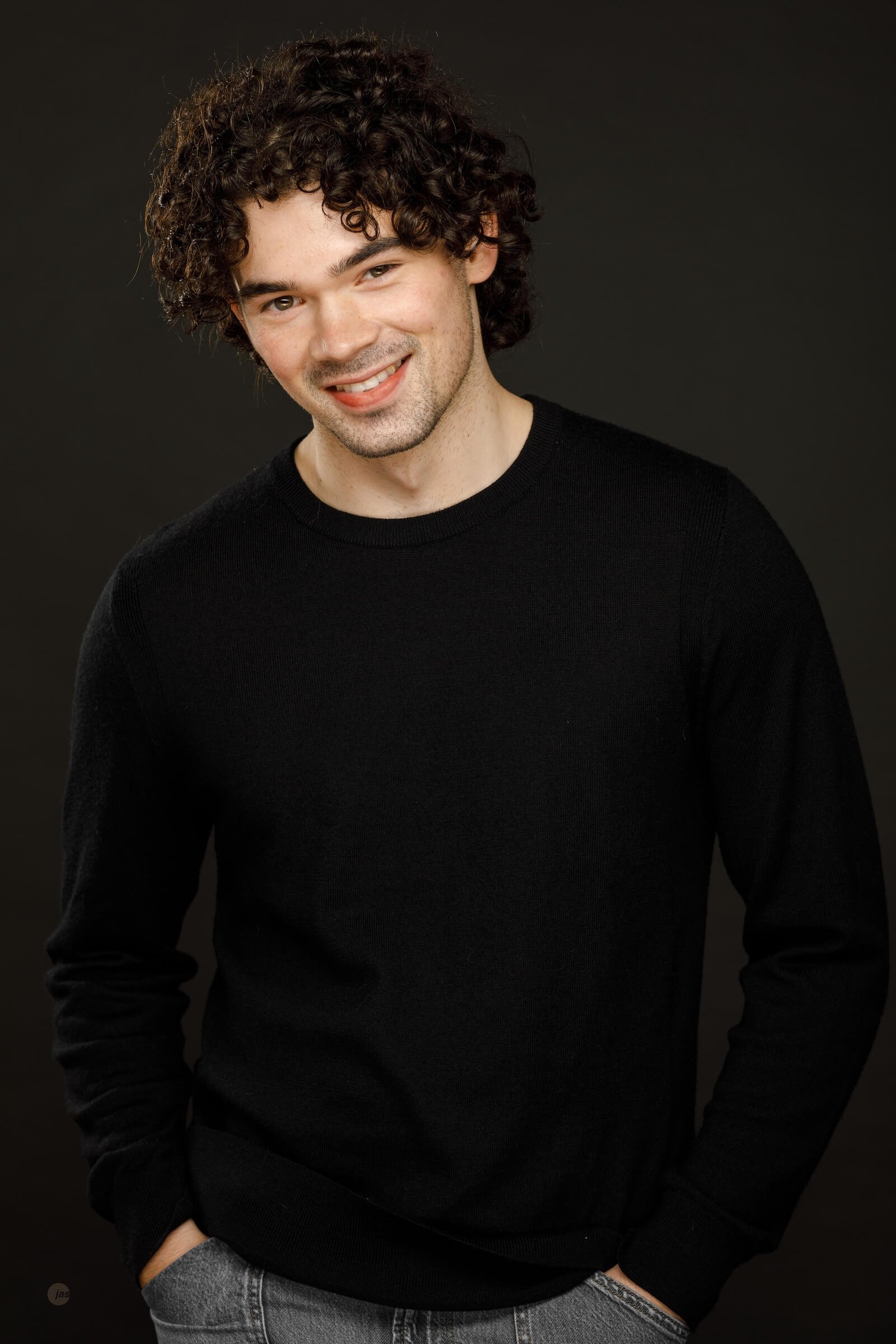 A young man with curly dark hair smiling, wearing a black sweater and gray pants, standing against a dark background.