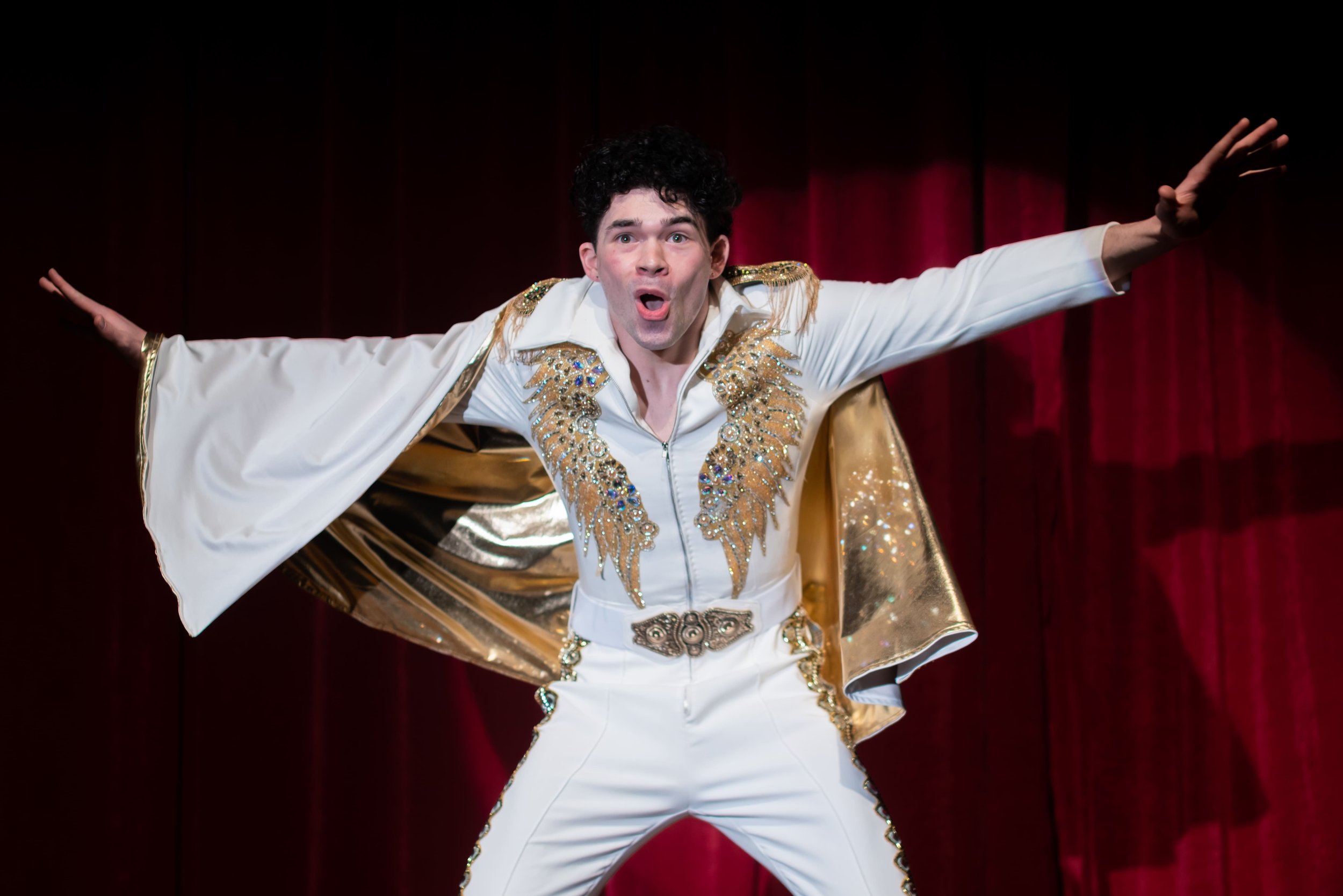 A man dressed as Elvis Presley in a white jumpsuit with gold and rhinestone embellishments, performing on stage with red curtains in the background.