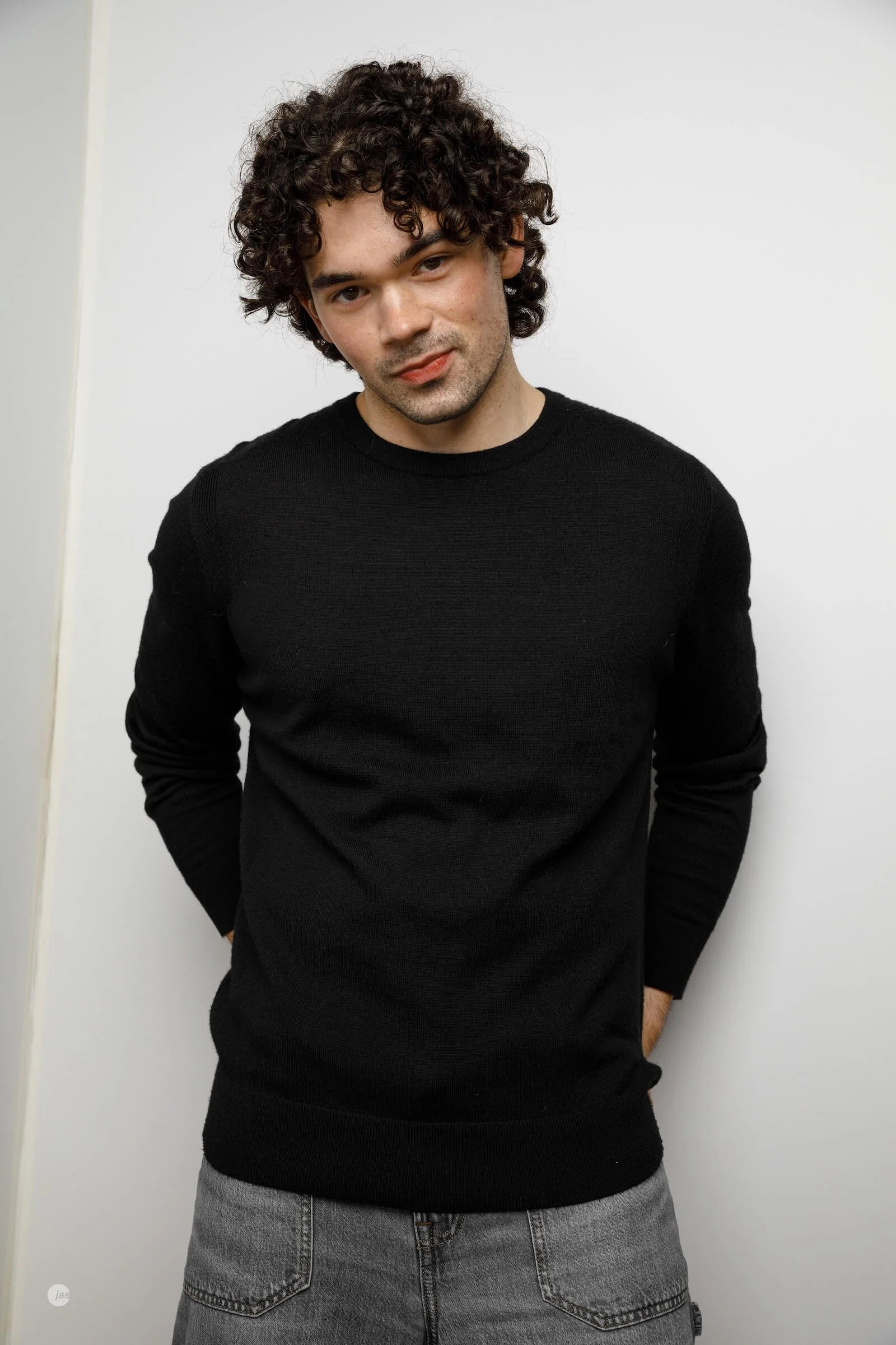 A young man with curly dark hair, light skin, and a slight beard, wearing a black sweater and gray jeans, standing against a plain white background with his hands in his pockets.