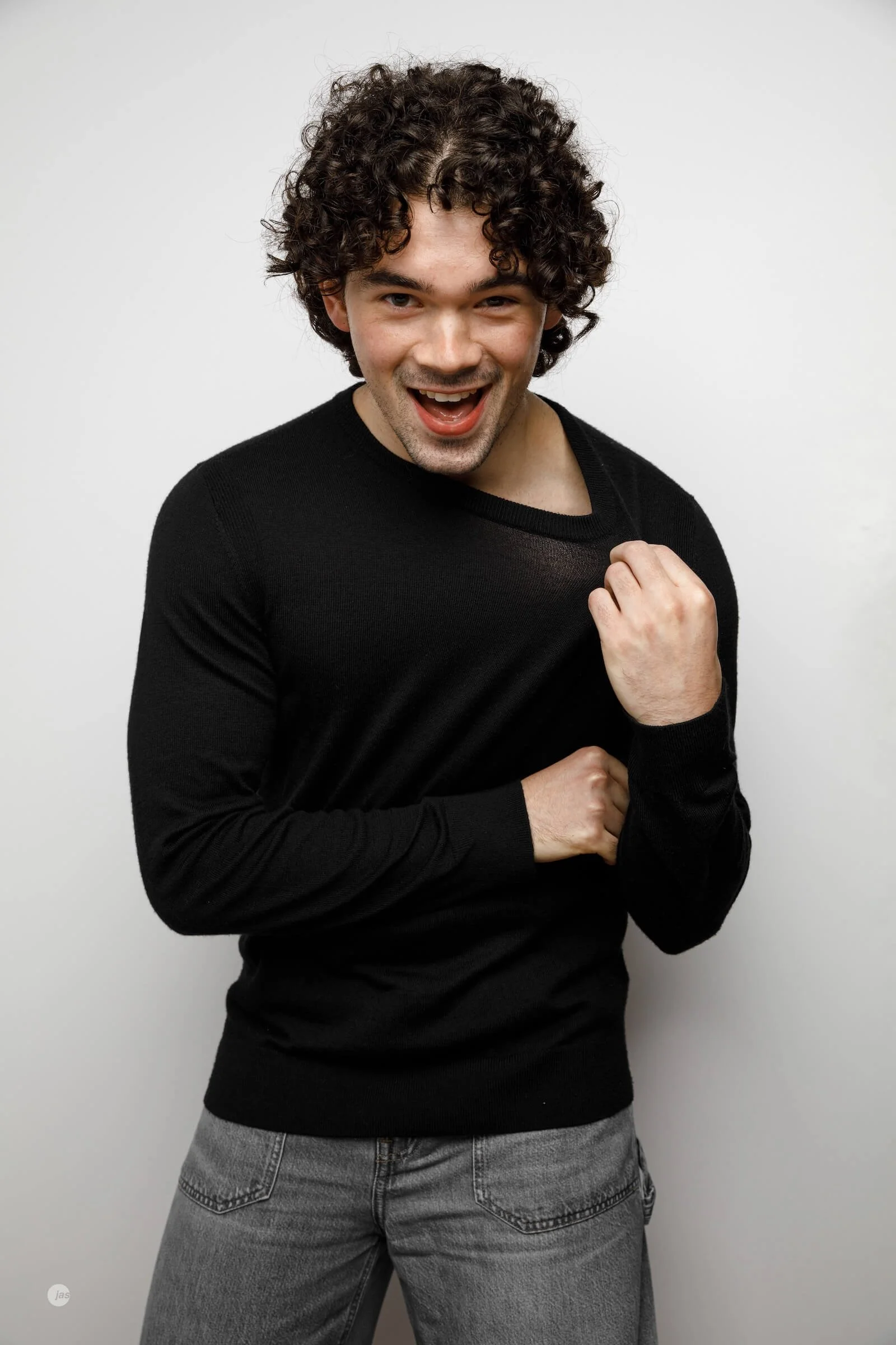 A young man with curly hair smiling and laughing, wearing a black sweater and gray jeans, standing against a plain white background.