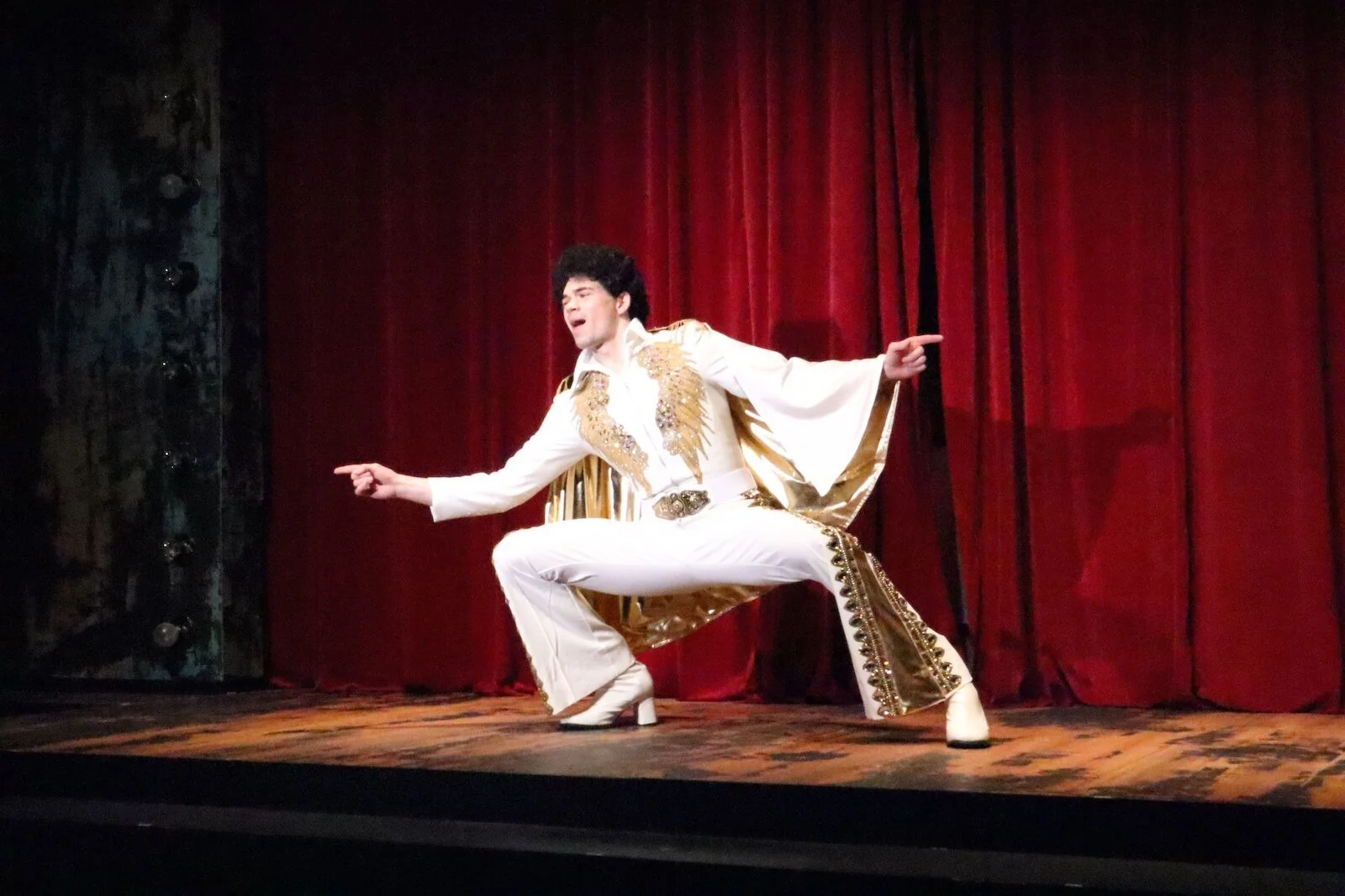 Performer dressed as Elvis Presley singing and dancing on stage with red curtains in the background.