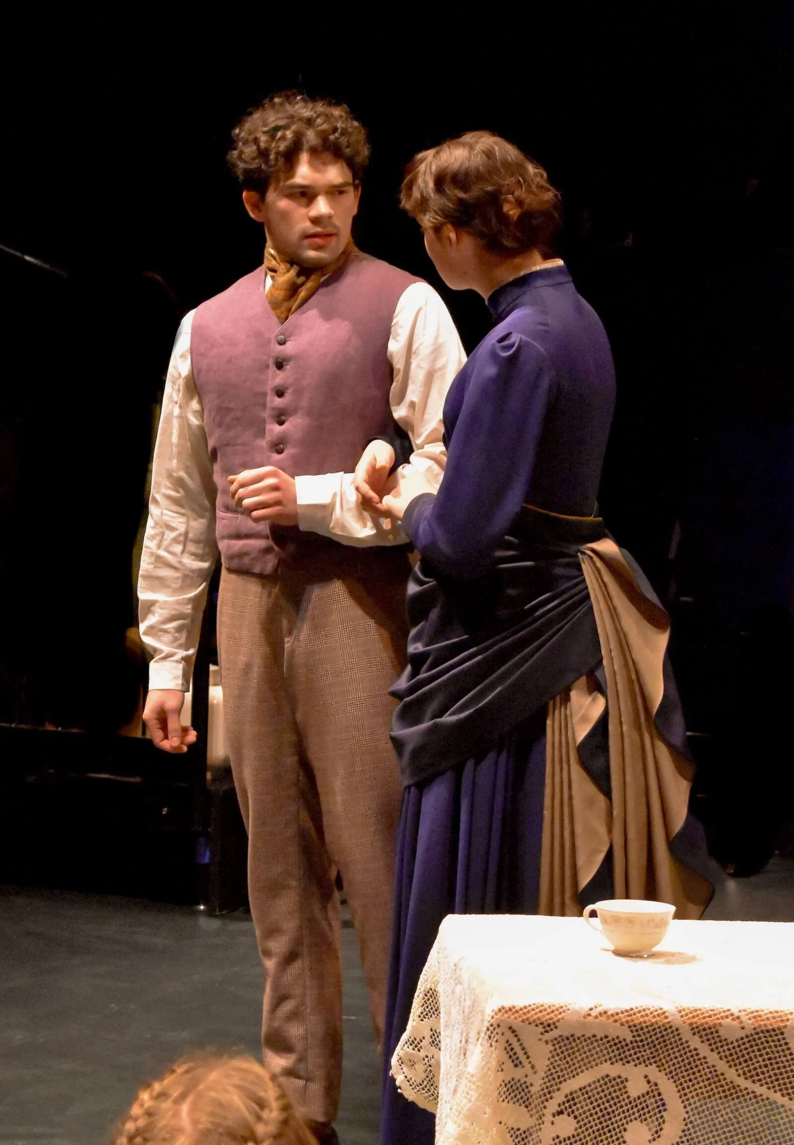 Two people in vintage costumes engaged in a scene on stage, with a small table and a teacup in front of them.