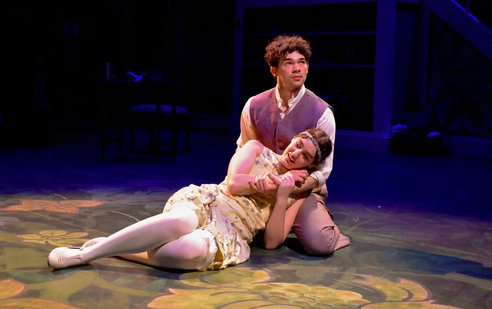Two actors performing on stage: a man sitting with a woman lying on his lap, both in vintage costumes. The woman appears to be emotionally expressive, while the man looks thoughtful. The stage has dark background with minimal set pieces.