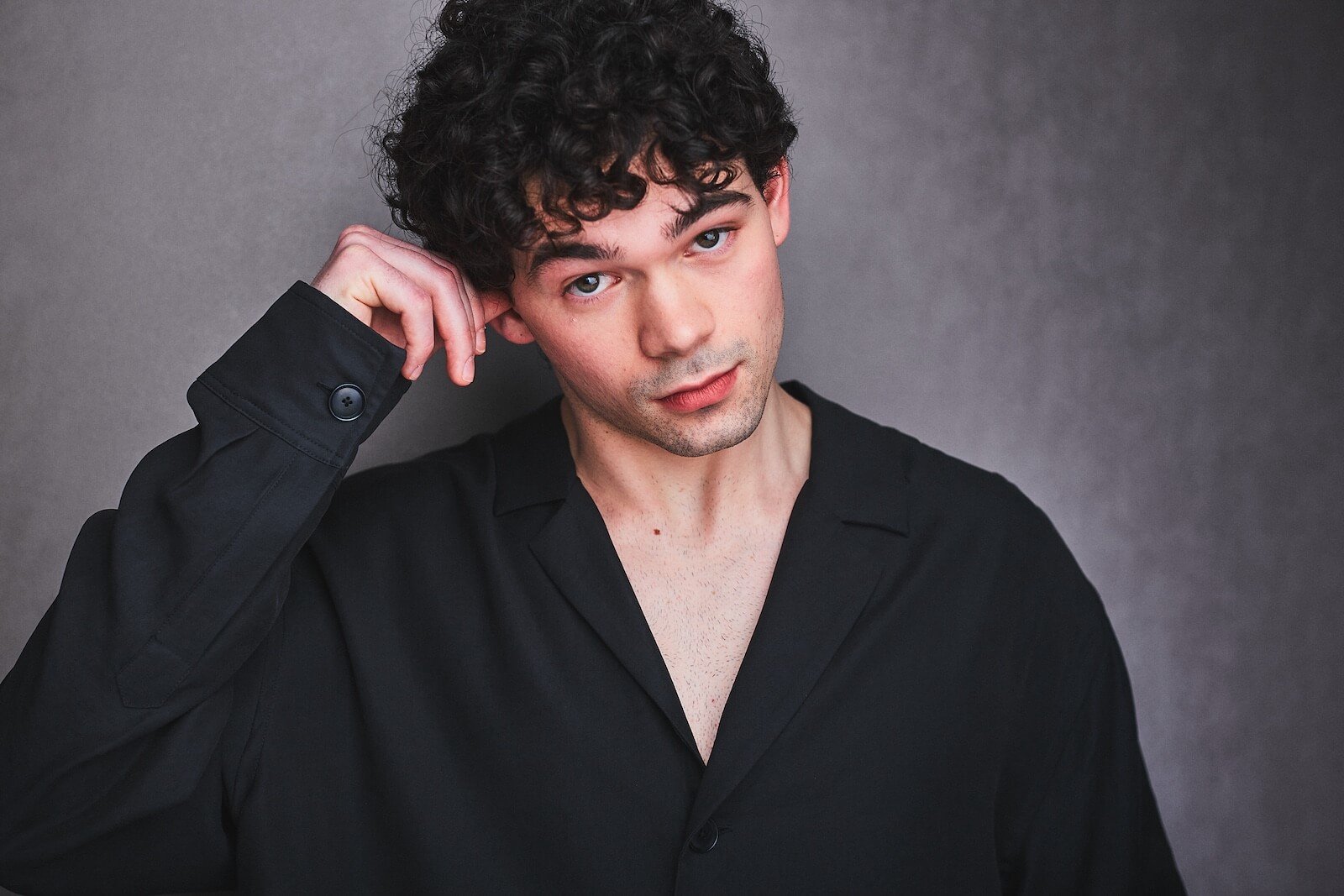 A young man with curly dark hair wearing a black shirt, resting his head on his hand, against a plain gray background.