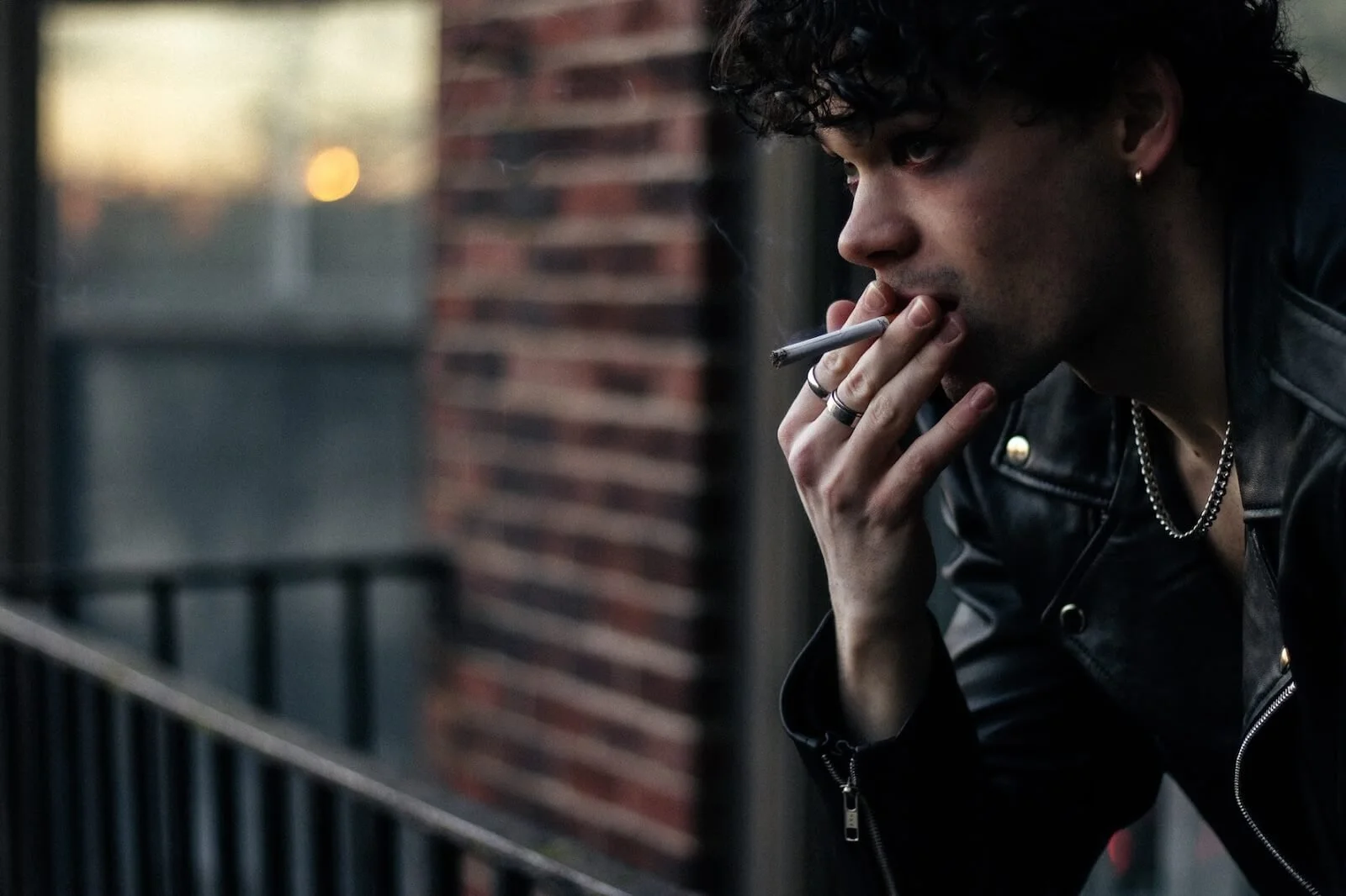 A young man with curly dark hair wearing a black leather jacket, silver rings, earrings, and a chain, smoking a cigarette on a balcony with a blurred cityscape in the background.
