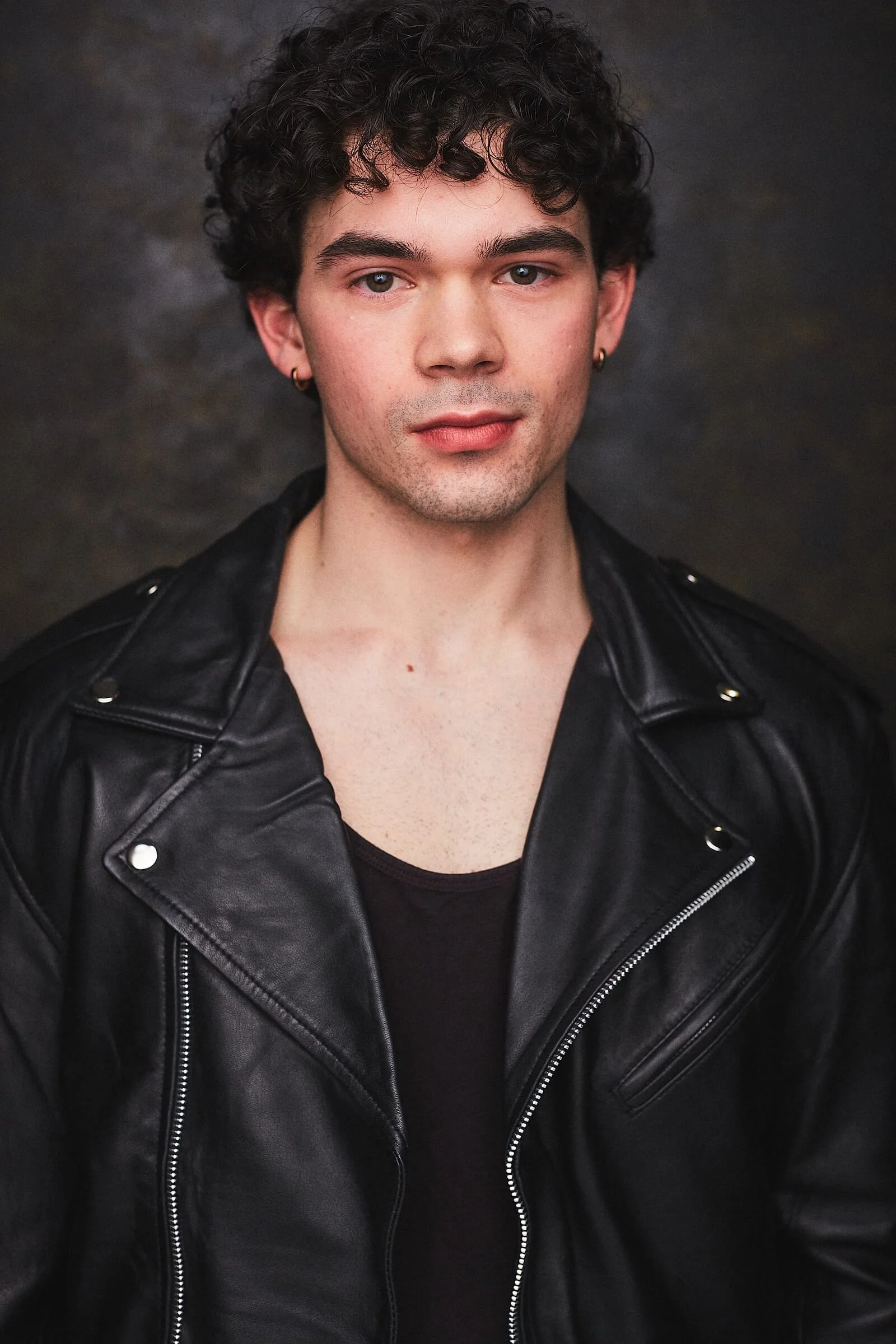 A young man with curly dark hair, light skin, and wearing a black leather jacket and earrings, posing against a dark background.
