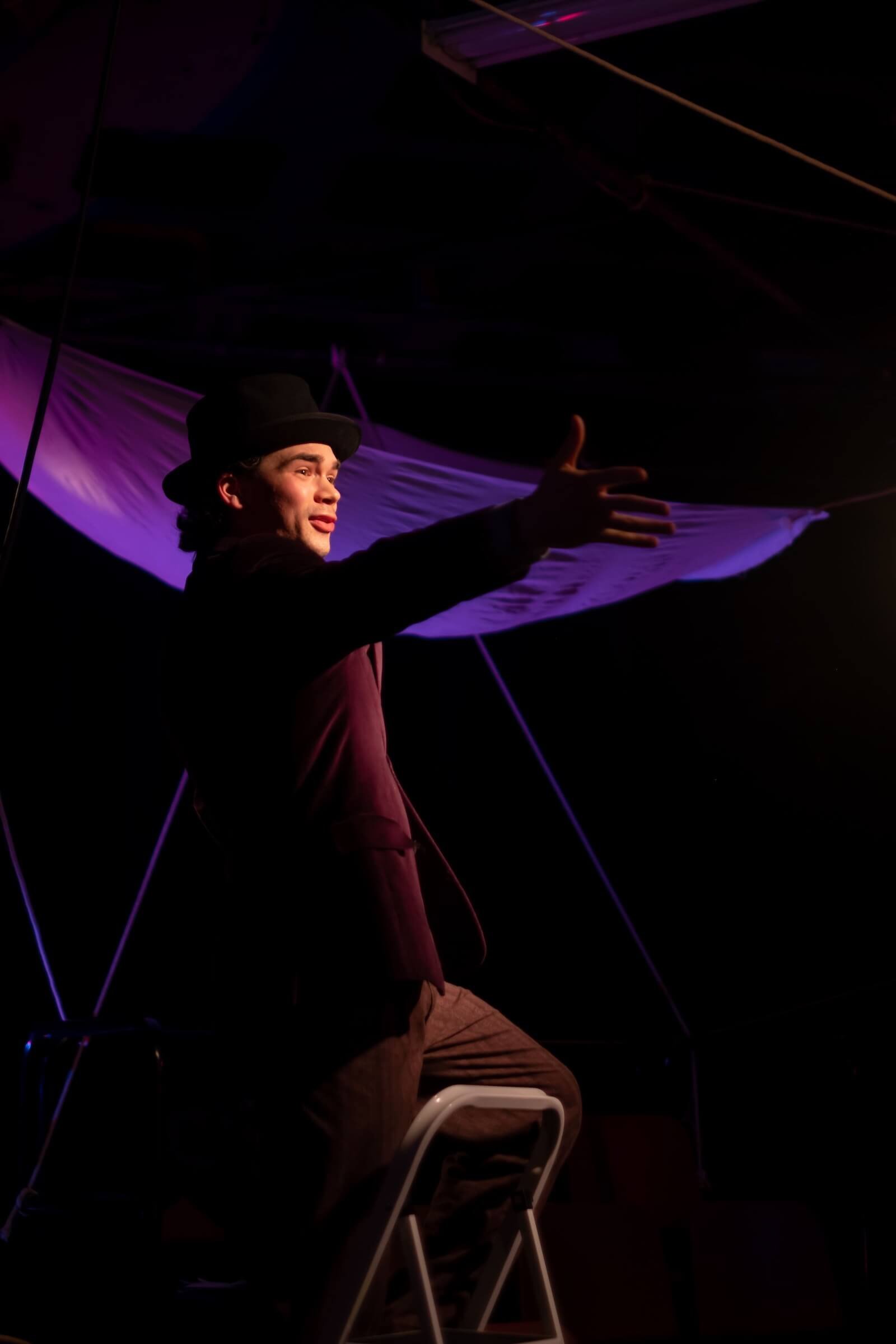 A person wearing a black hat and dark clothing, sitting on a white chair, engaged in a theatrical performance with purple lighting and fabric drapery in the background.