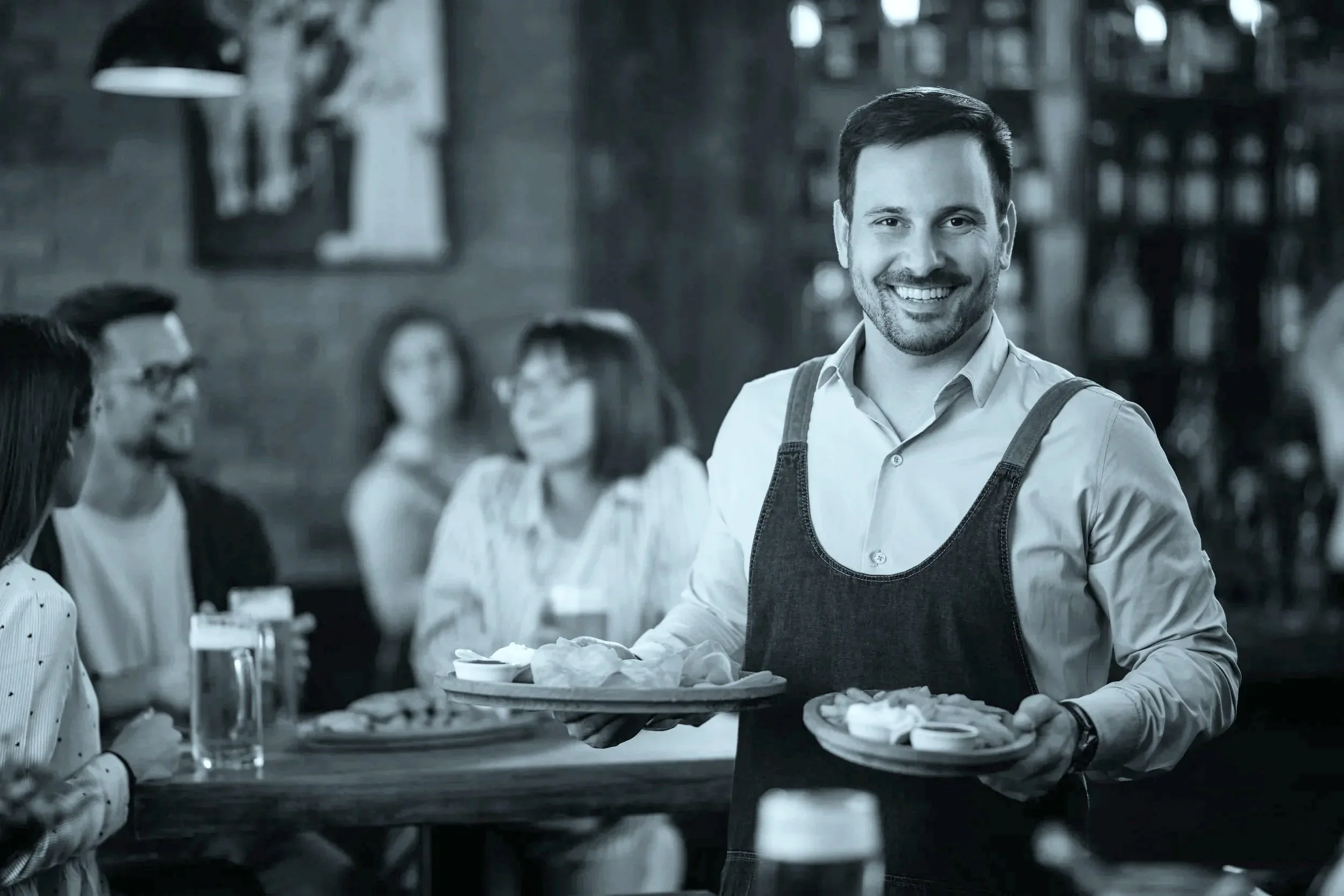 En smilende bartender bærer fade med appetizers i en travl hyggelig bar eller restaurant med flere gæster, der nyder drikkevarer.