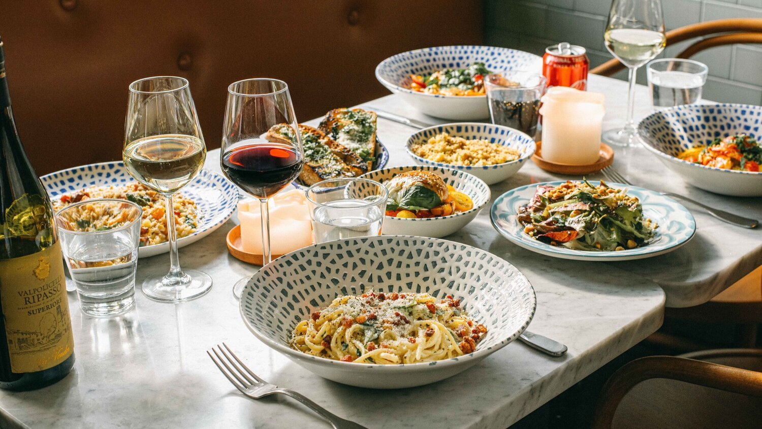 En bordstabel med flere fade af pasta, salat, grøntsager og brød, samt glas med hvidvin, rødvin og vand, en vinflaske og en tændt stearinlys.