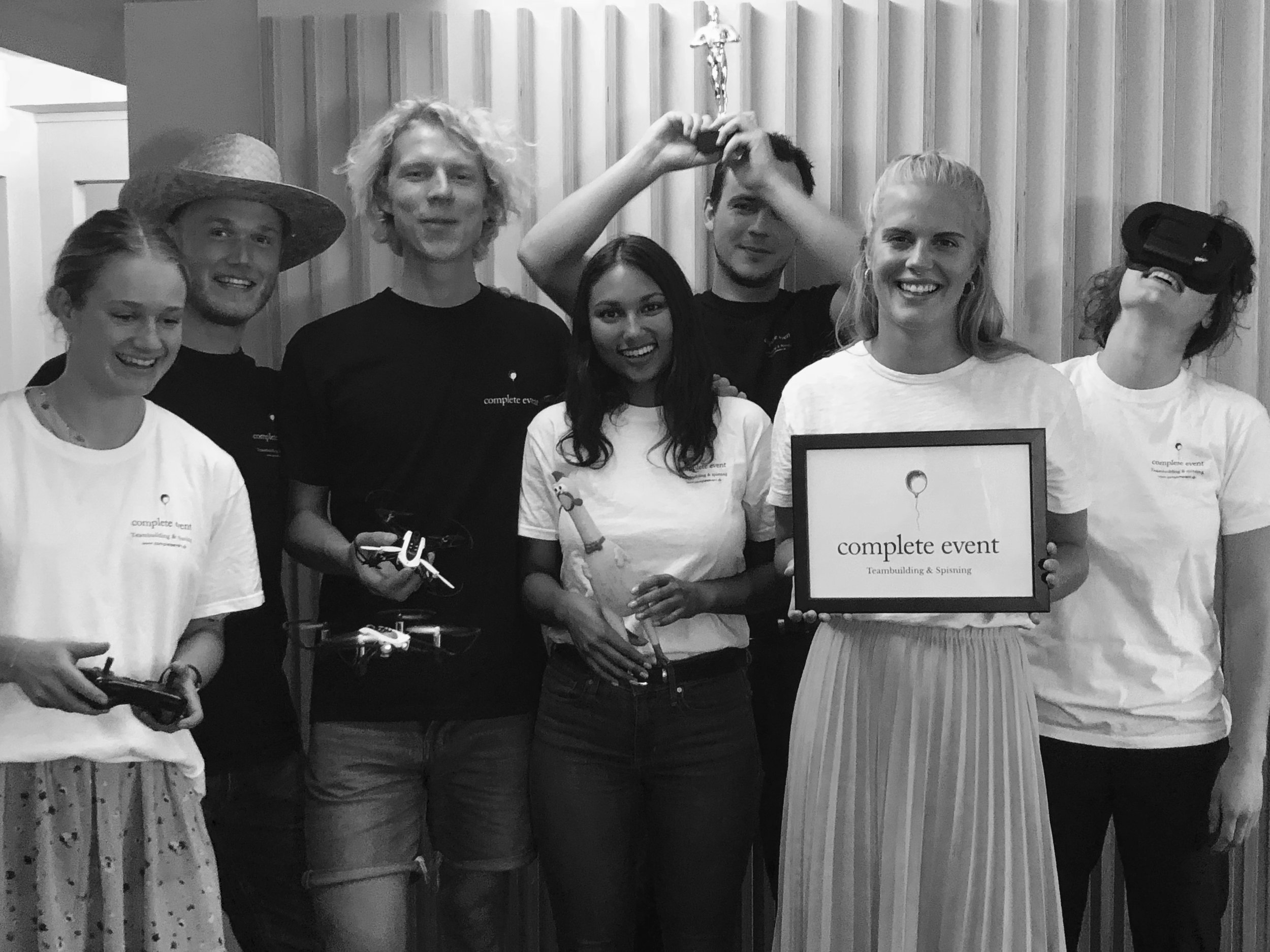 En gruppe mennesker smiler, nogle holder fjernbetjeninger, en person har VR-briller på, og en kvinde holder en skærm med teksten 'complete event'.