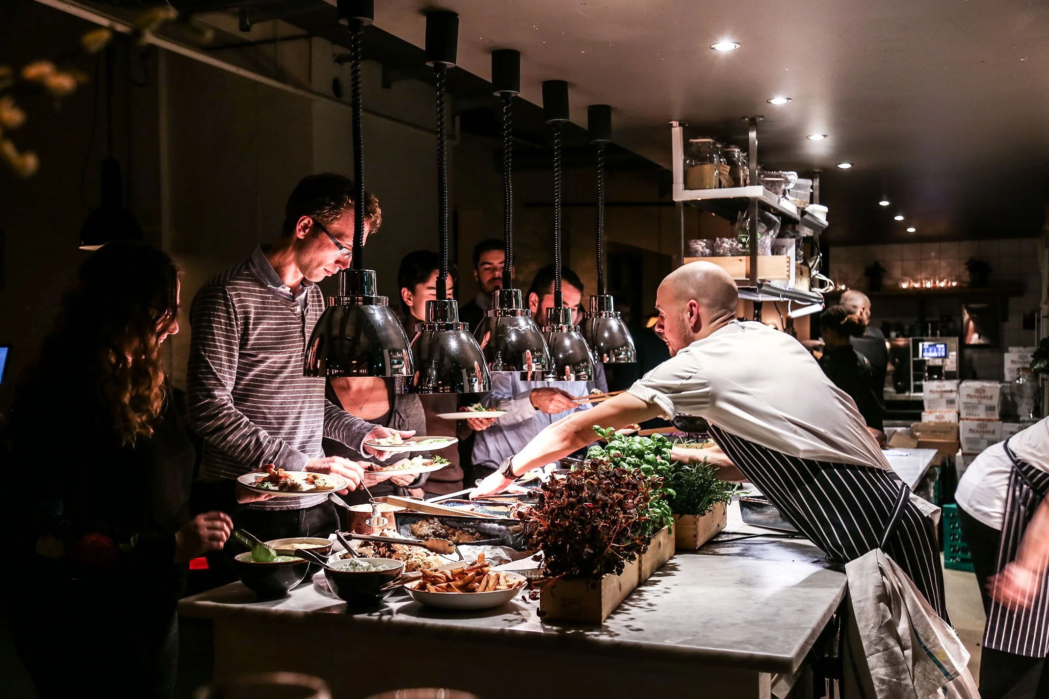 Konkret et buffet ved et restaurant, hvor en kok serverer mad til gæster, der venter i kø, omgivet af lamper og køkkenudstyr.