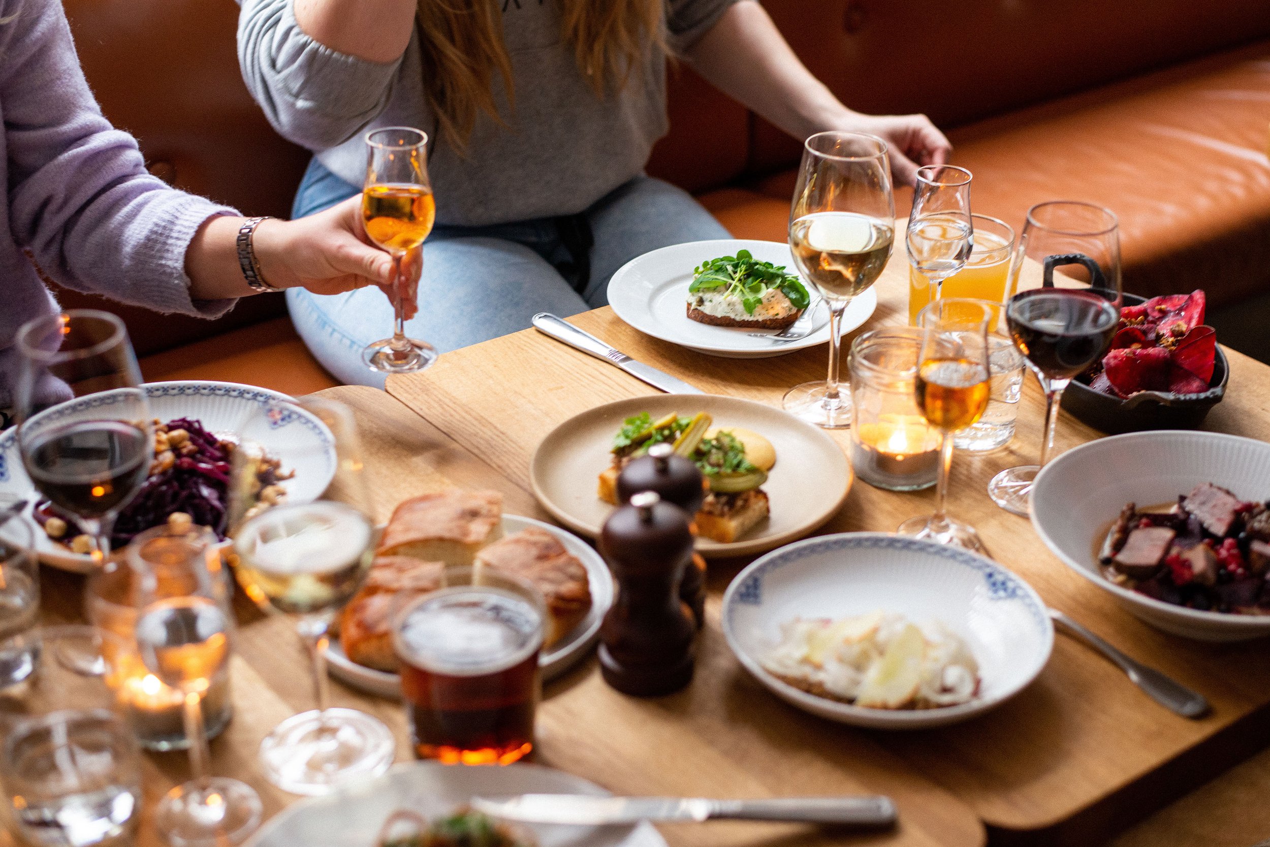 Et bord med mad og drikkevarer til en middag, inklusive glas vin, øl, og forskellige retter som salater, kød og brød, omgivet af skærebrætter og lys.