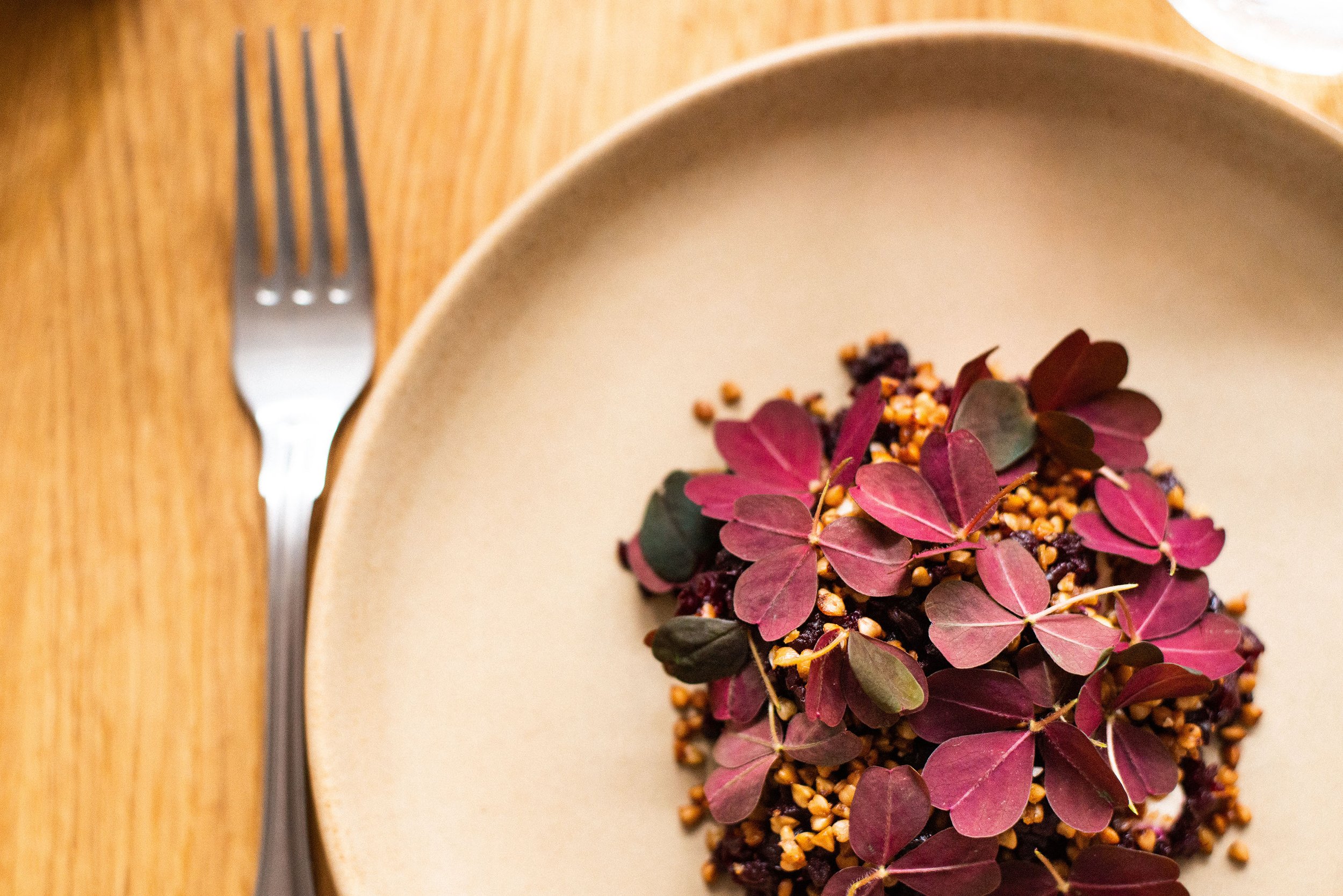 En tallerken med en madret med mørkeblade og små, gule og sorte ingredienser. Det er en beige tallerken og en gaffel til venstre på et træt bord.