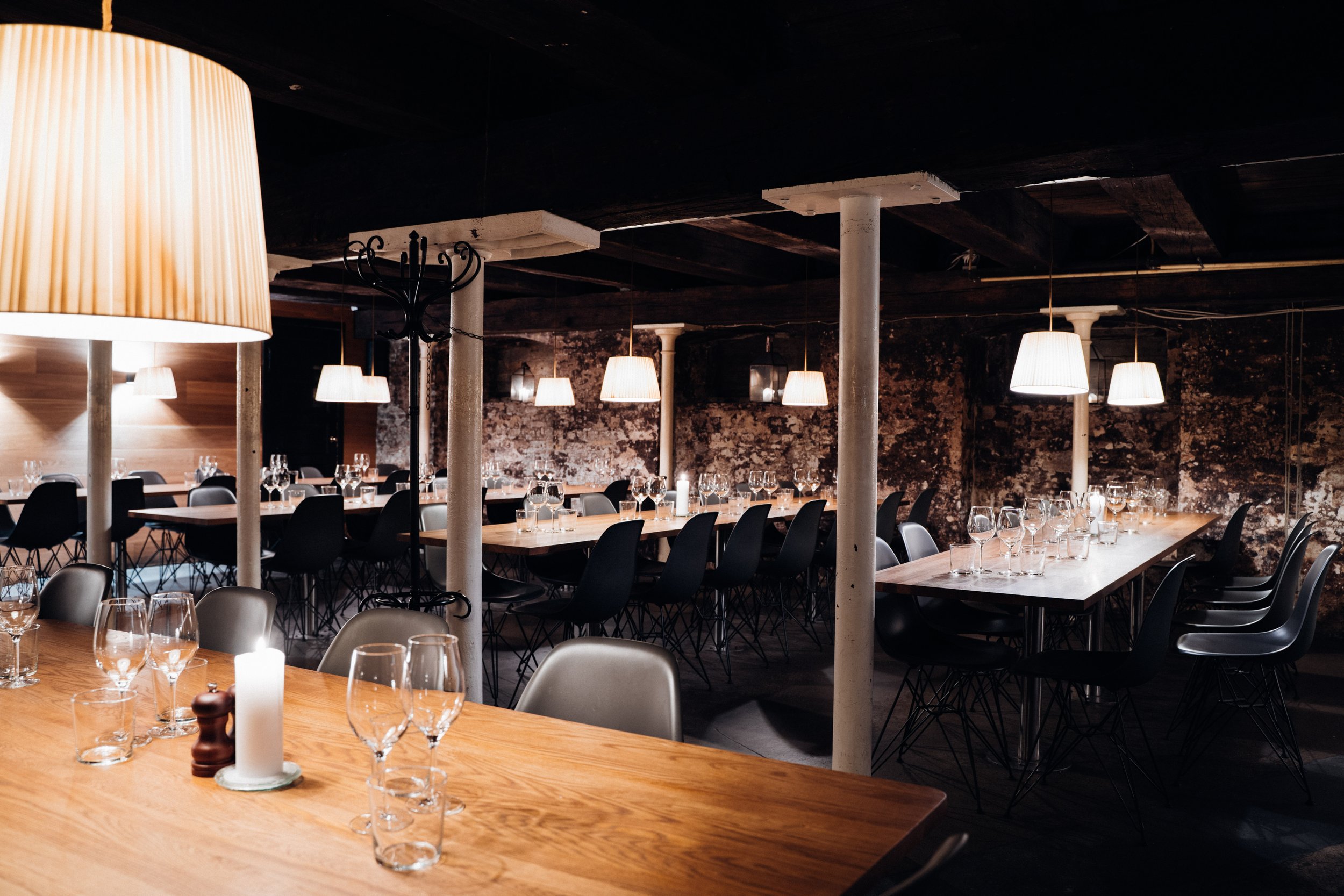 Indrettet restaurant med bord dækket med glas og bestik, moderne lamper, rustik vægbaggrund og stole omkring bordene.