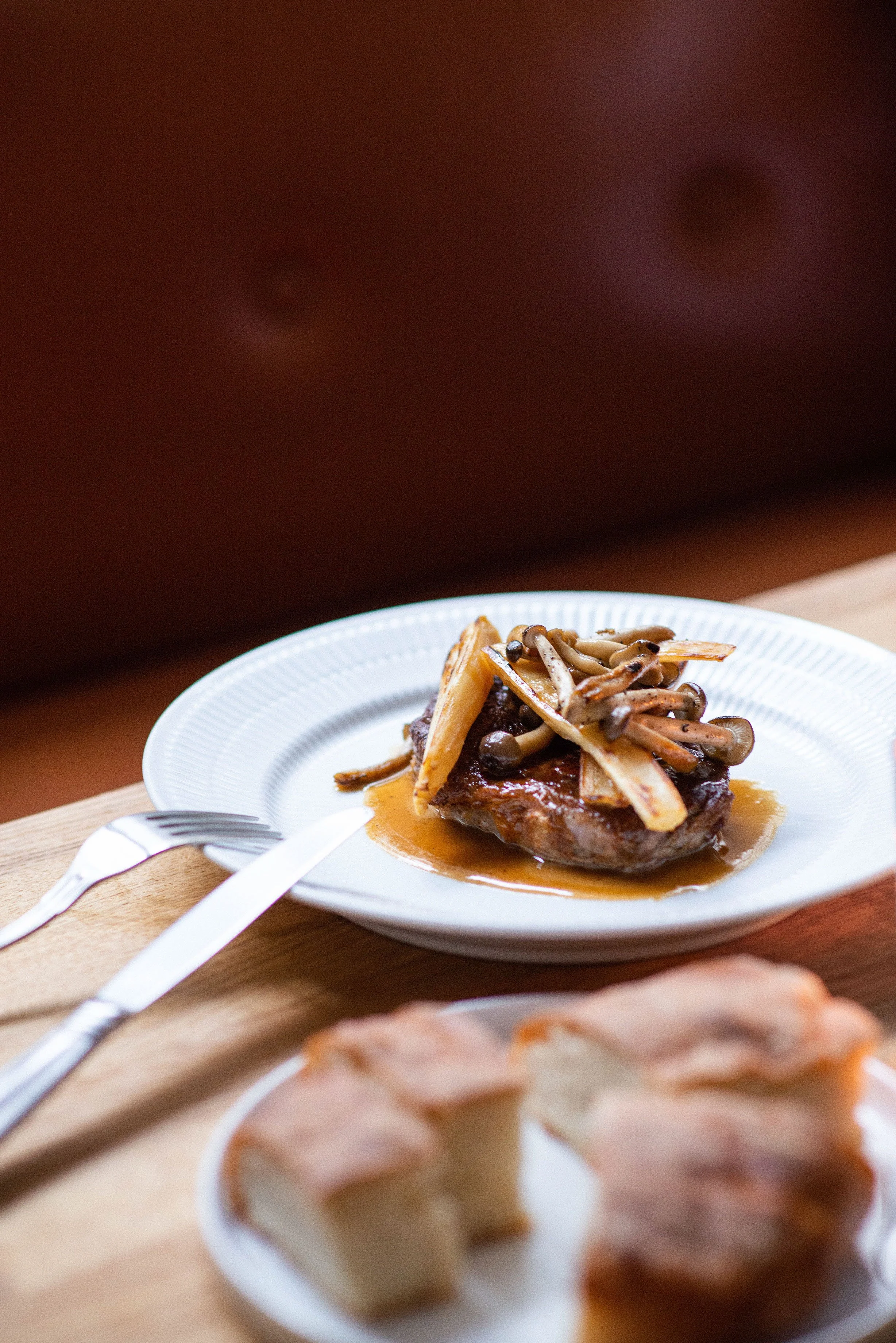 En tallerken med bøf toppet med stegte champignoner og pommes frites, serveret med sovs. Et stykke af anden ret i baggrunden.