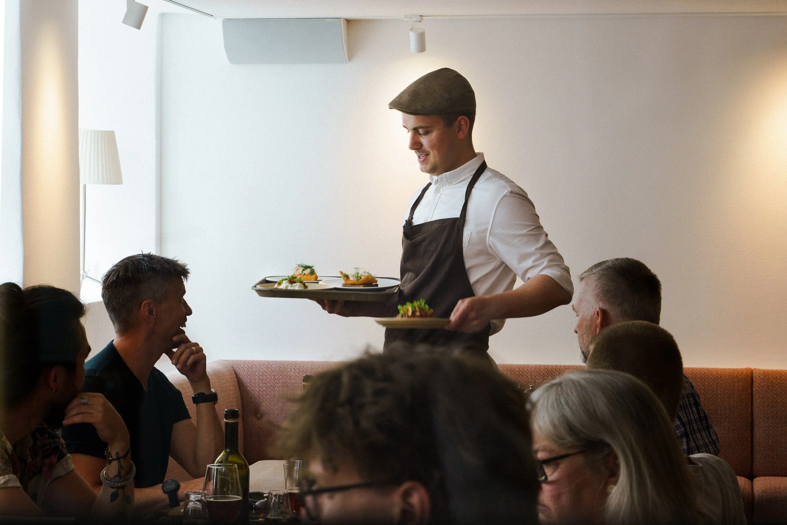 En tjener serverer små retter ved et bord i en restaurant, mens gæster sidder og nyder maden.