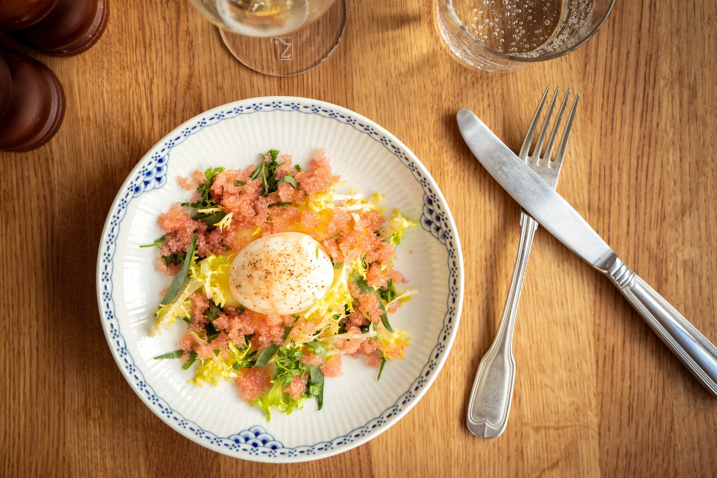 En tallerken med en salat med æg, grønne blade og pink kaviar. Serviet, kniv og gaffel på et træbord. Drikkevarer i baggrunden.