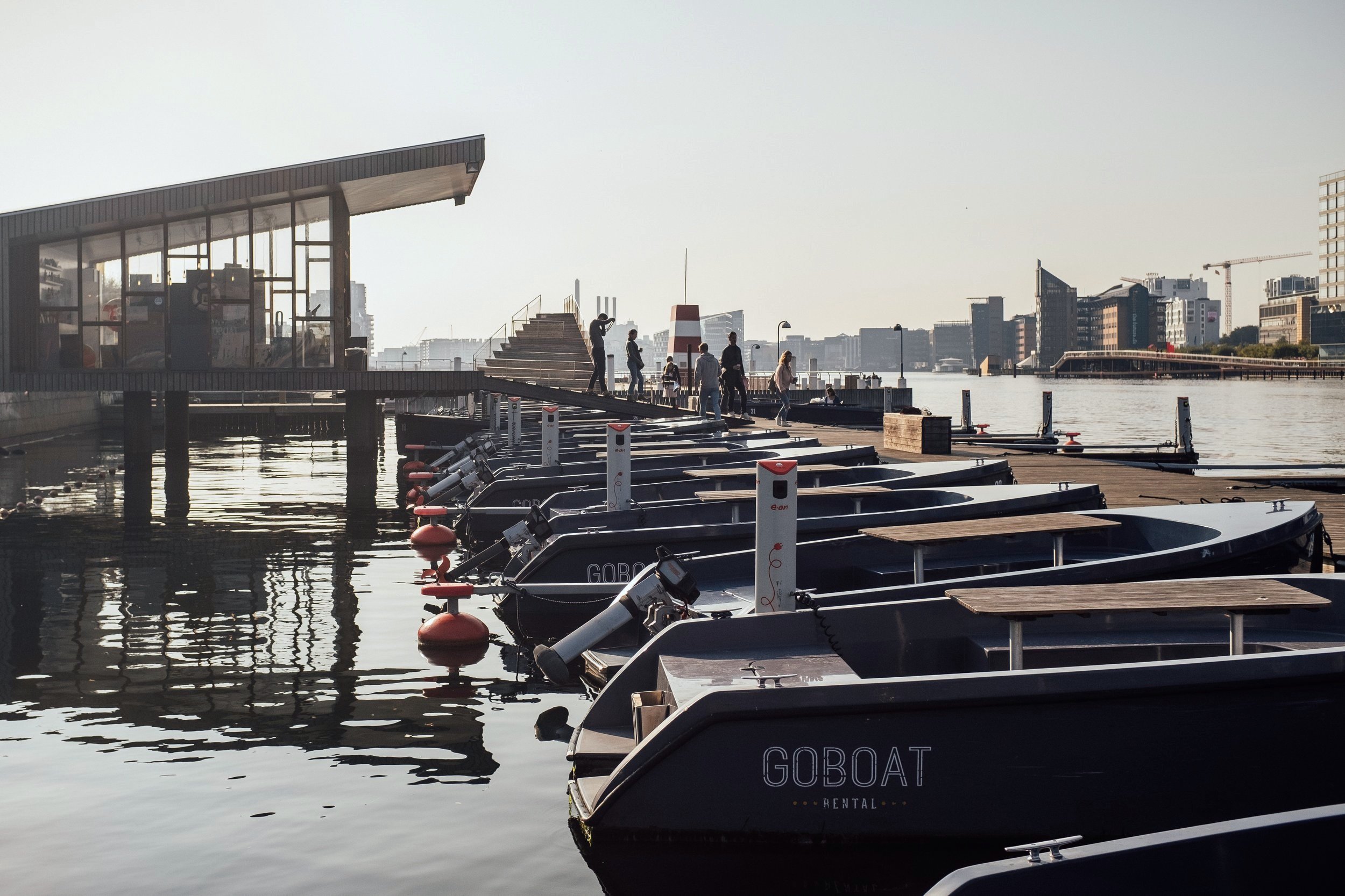 Bådbro med flere små både ved kajen, mennesker gående på dækket, moderne bygninger i baggrunden, og solnedgang eller morgenlys.