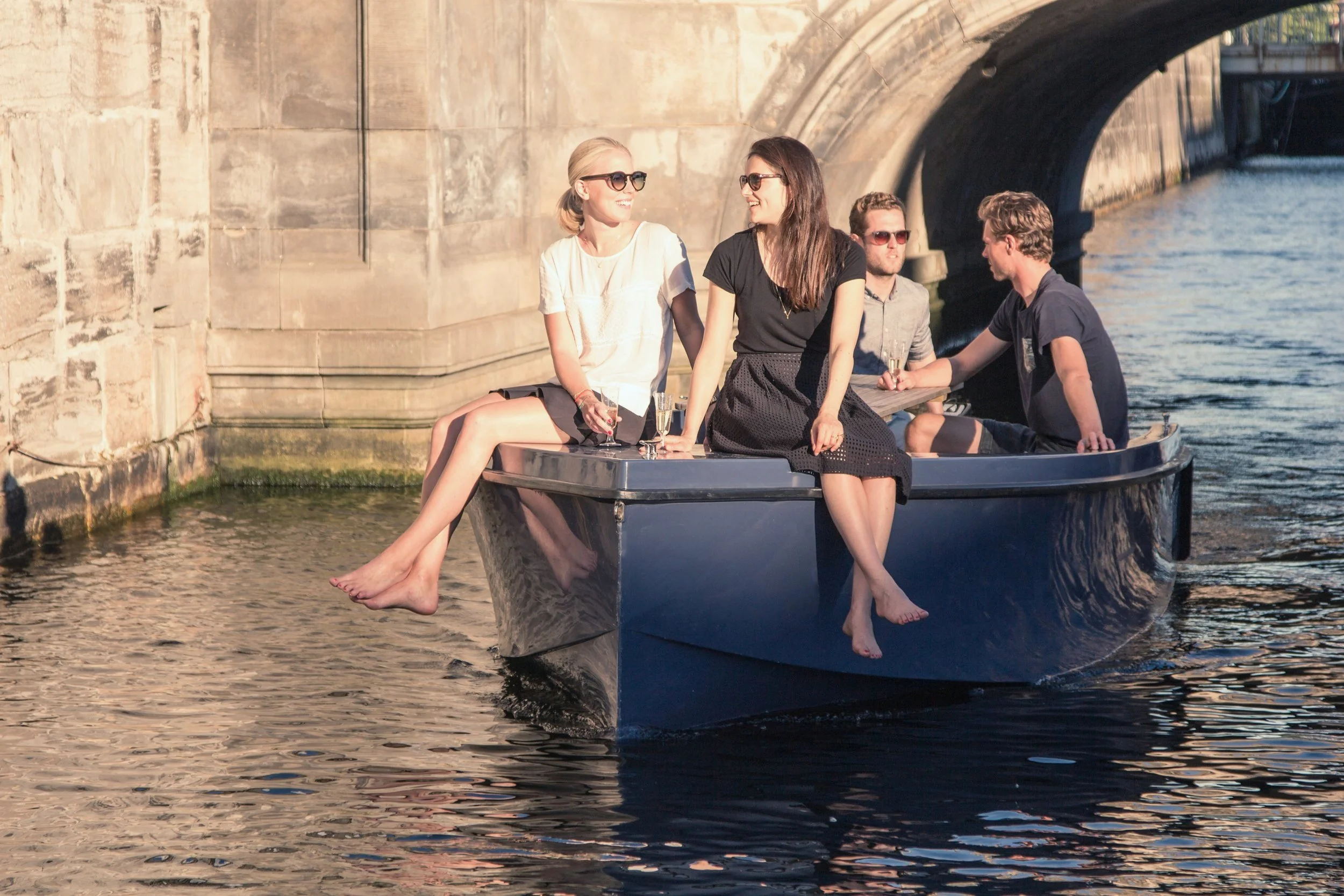 Fire voksne og to børn, sidder i en båd under en bro på en flod i solskin, drikker champagne og nyder det gode vejr.