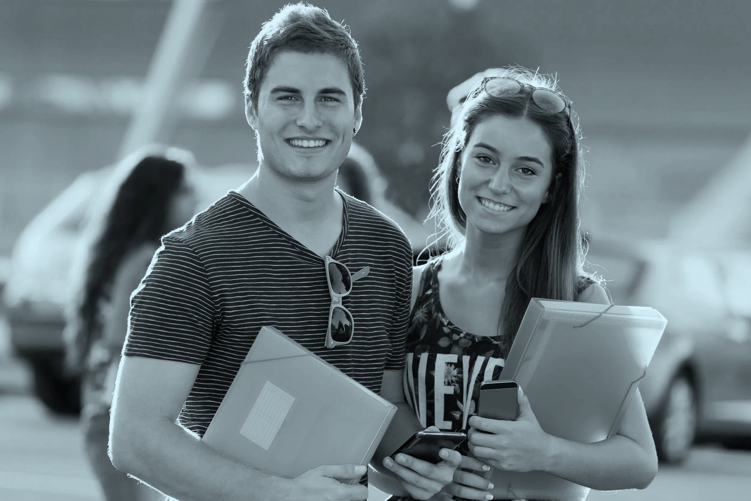 To unge mennesker, en mand og en kvinde, smiler med skolobøger og smartphones i hænderne. De står udenfor, med biler i baggrunden.