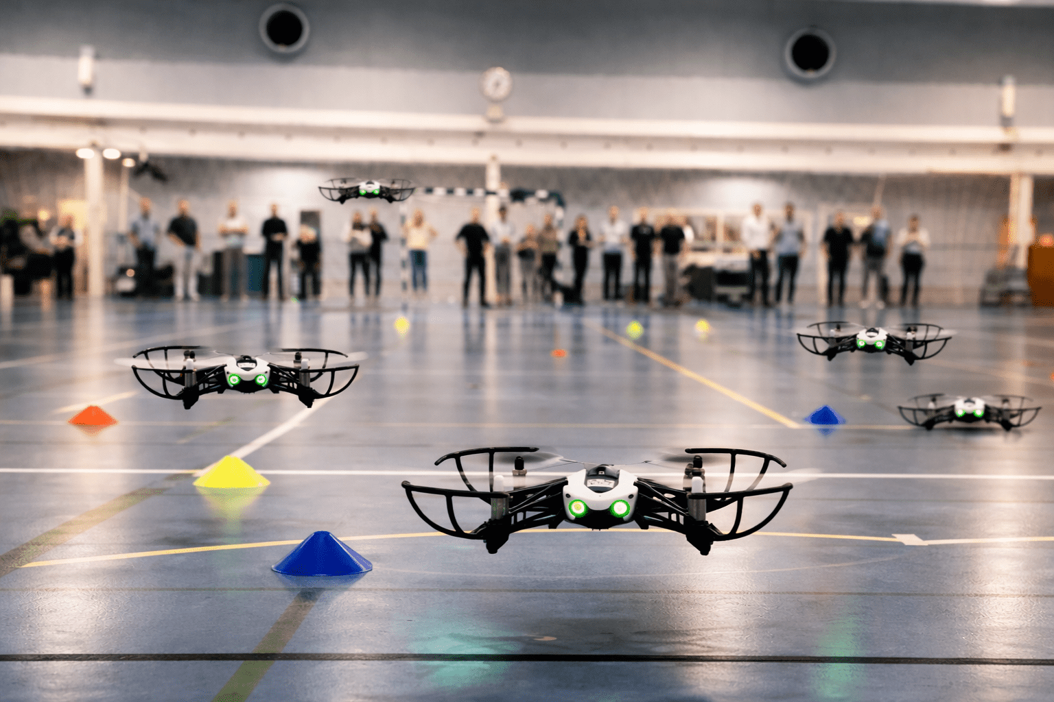 Fire droner flyver i en sportshal med 3 svømmende droner i forgrunden. Der er farvede kegler på gulvet og en gruppe mennesker står bag i baggrunden.