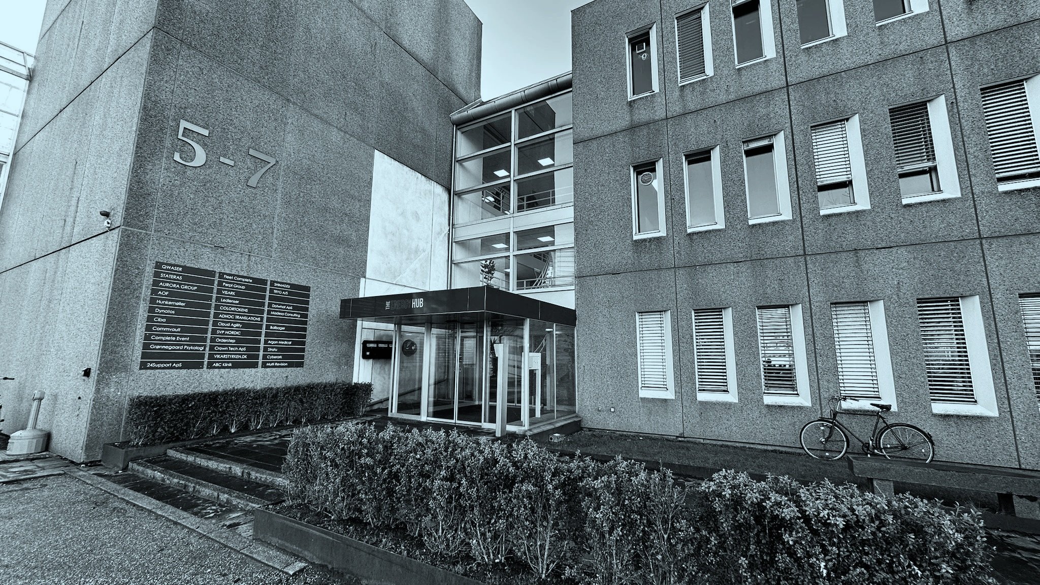 Uden for en kontorbygning med adressen 5-7, en sort indgang med glassdøre, en cykel parkeret mod væggen, og en sort tavle med samarbejdspartnere på væggen