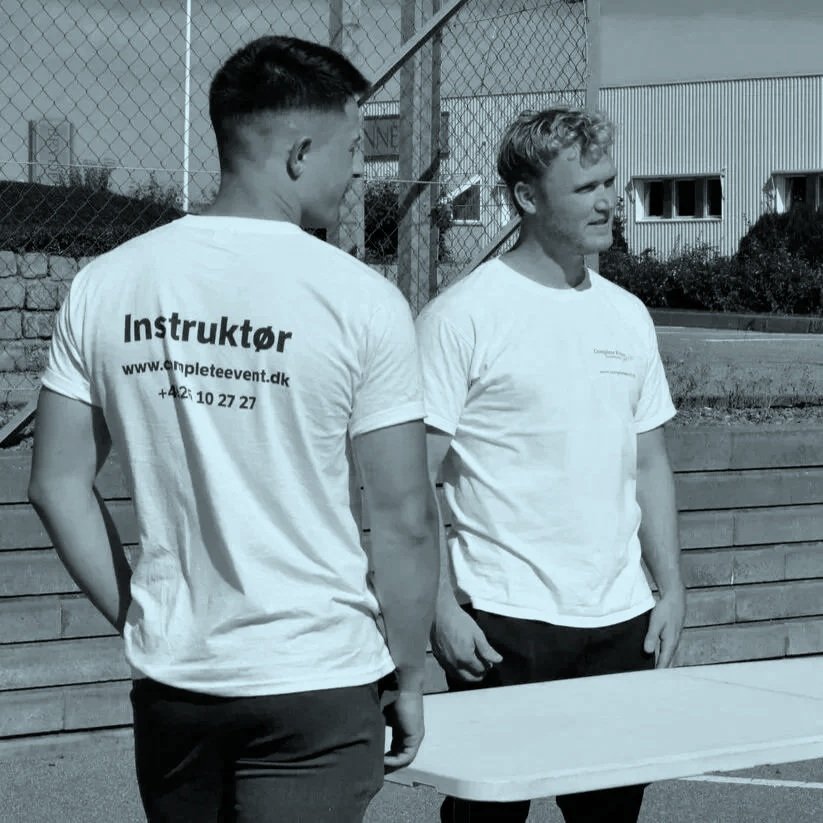 To unge mænd står overfor hinanden udenfor, iført hvide T-shirts og taler, med en bord foran dem. En af dem har teksten 'Instruktør' på ryggen.