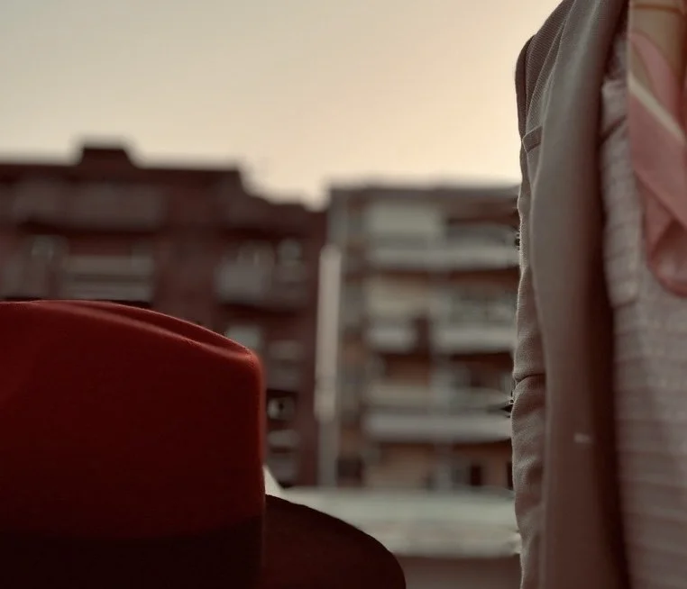 Close-up shot of a red hat on a surface with a person's arm and torso partially visible on the right side, blurred building in the background.