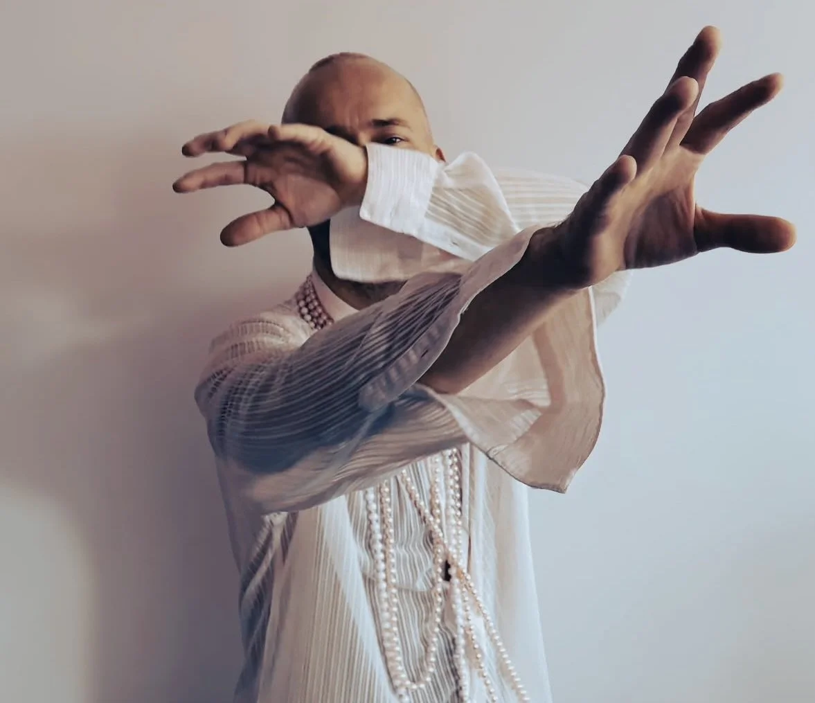 Person with a bald head wearing a white face mask and a pearl necklace, crossing their arms in front of them, dressed in a beige, pleated, sheer top, against a plain light background.