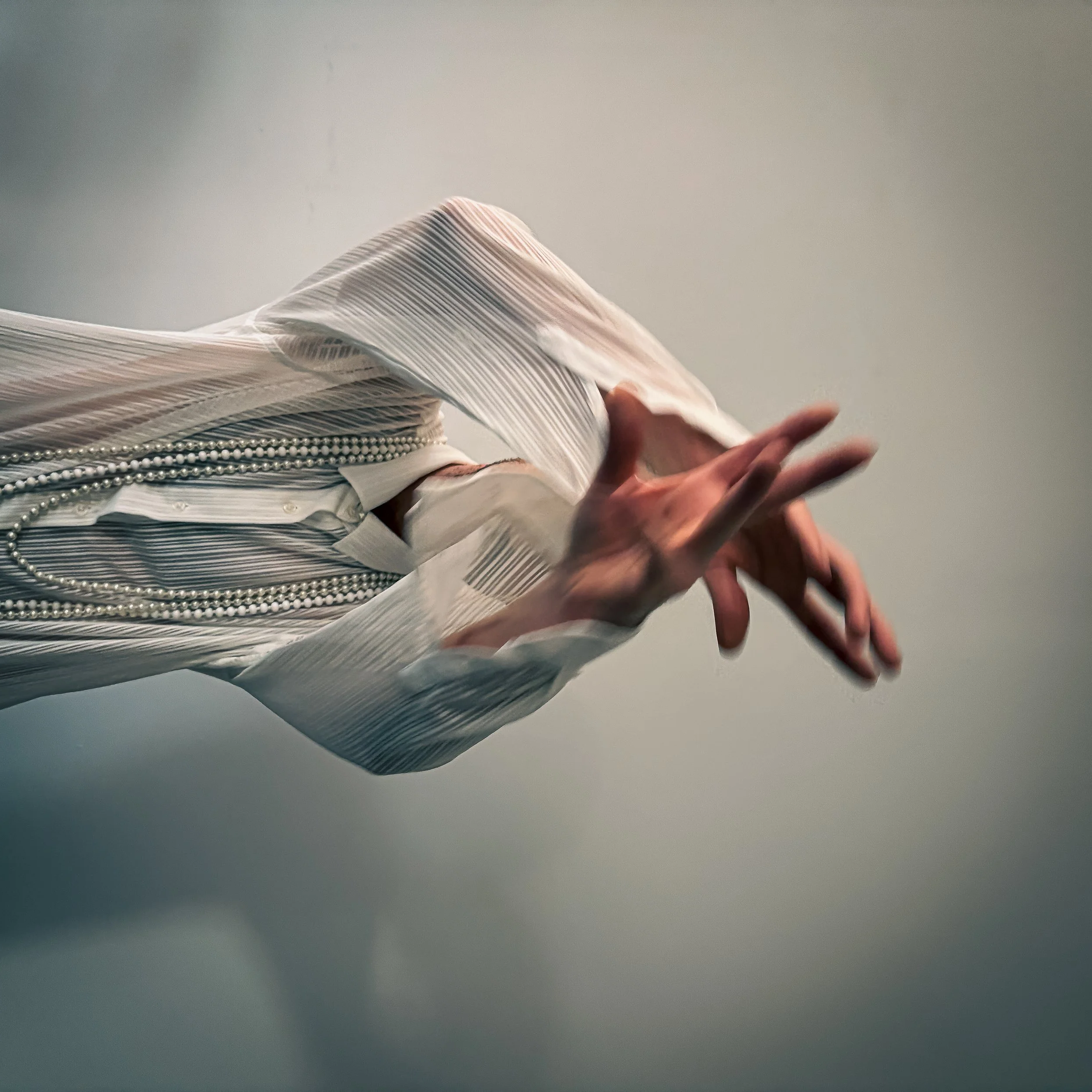 Close-up of a person wearing a white pleated shirt and pearl necklaces, with their hand gesture in front of a plain background.