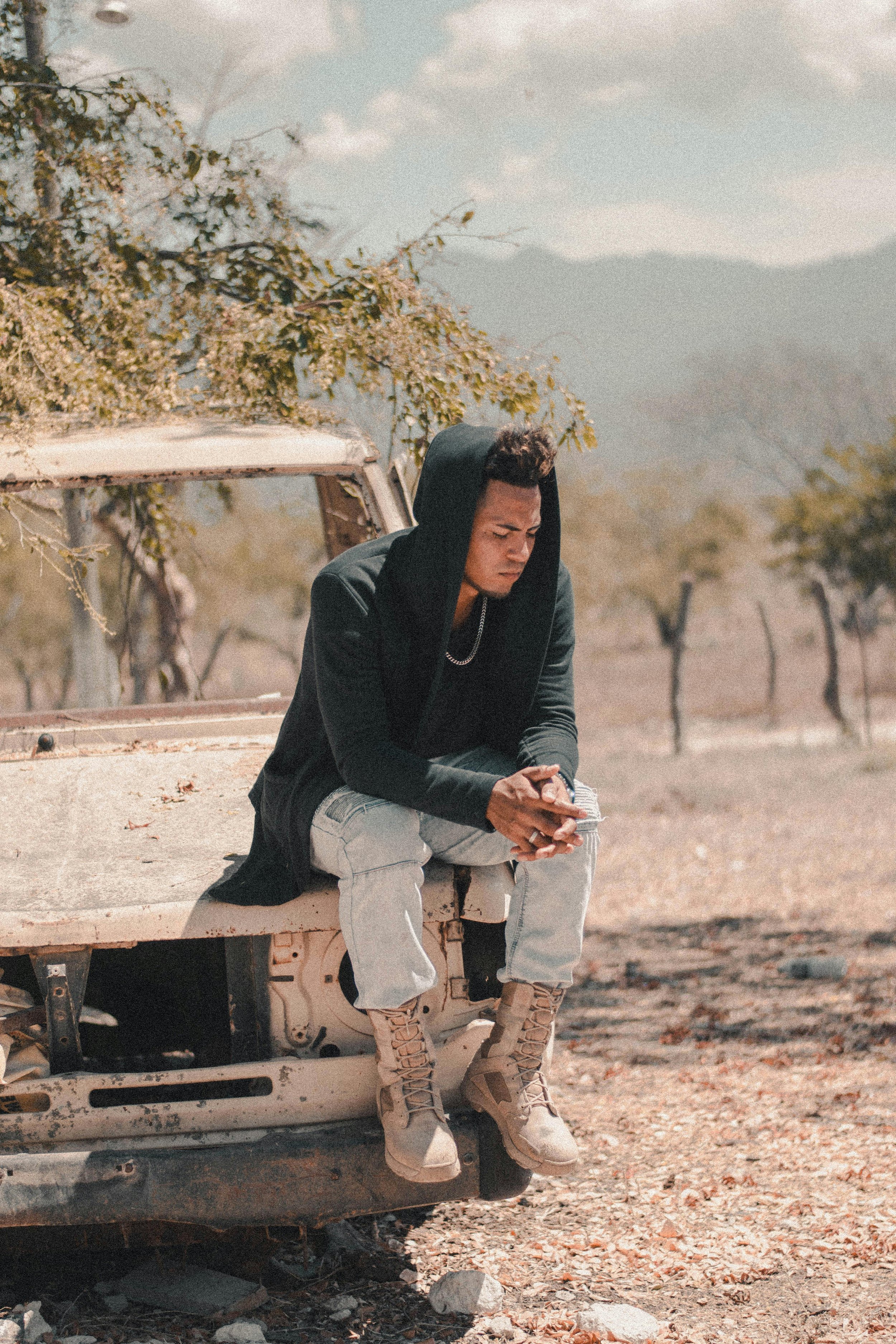 A young man with a hoodie, chain, light jeans, and beige boots sitting on the edge of an old, rusted truck in a rural area with dry land and trees, looking at his phone.