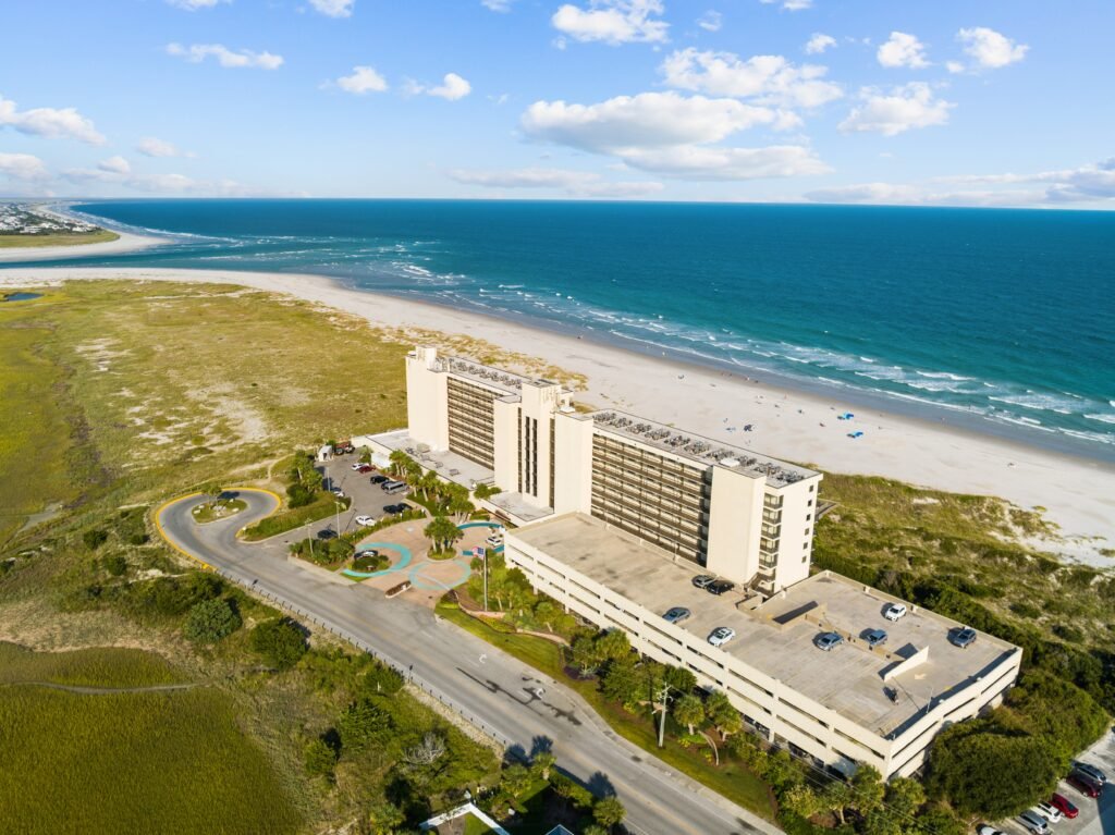 Aerial view of a beachfront hotel next to a sandy beach, with blue ocean waves and partly cloudy sky.