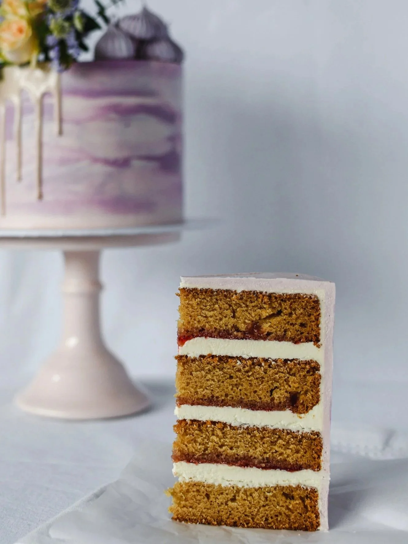 A slice of layered cake with white icing and a visible fruit filling, set on a white surface with a blurred purple and white cake in the background.
