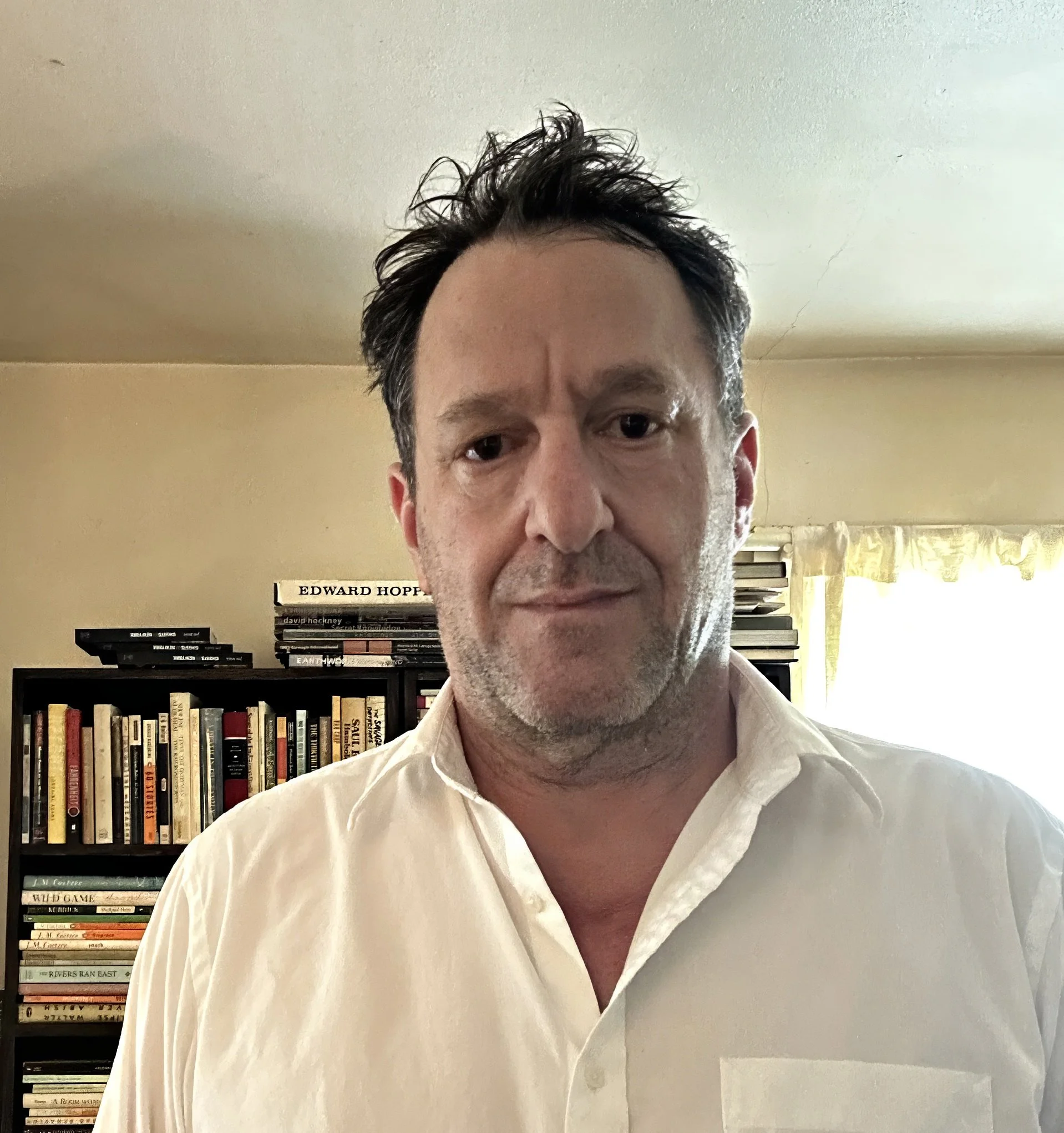 A middle-aged man with dark, slightly messy hair, wearing a white shirt, standing in a room with a bookshelf filled with books behind him and a window with yellow curtains.